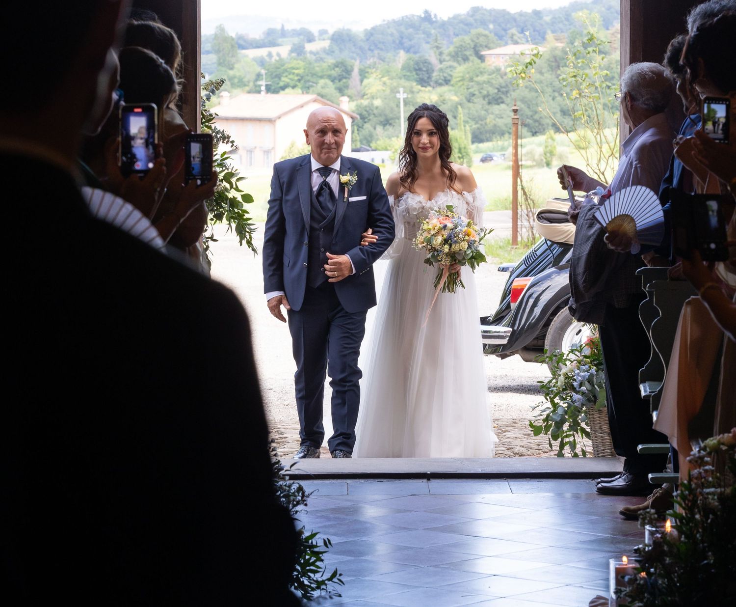 Wedding ceremony aisle with guests seated in white chairs under natural outdoor lighting.