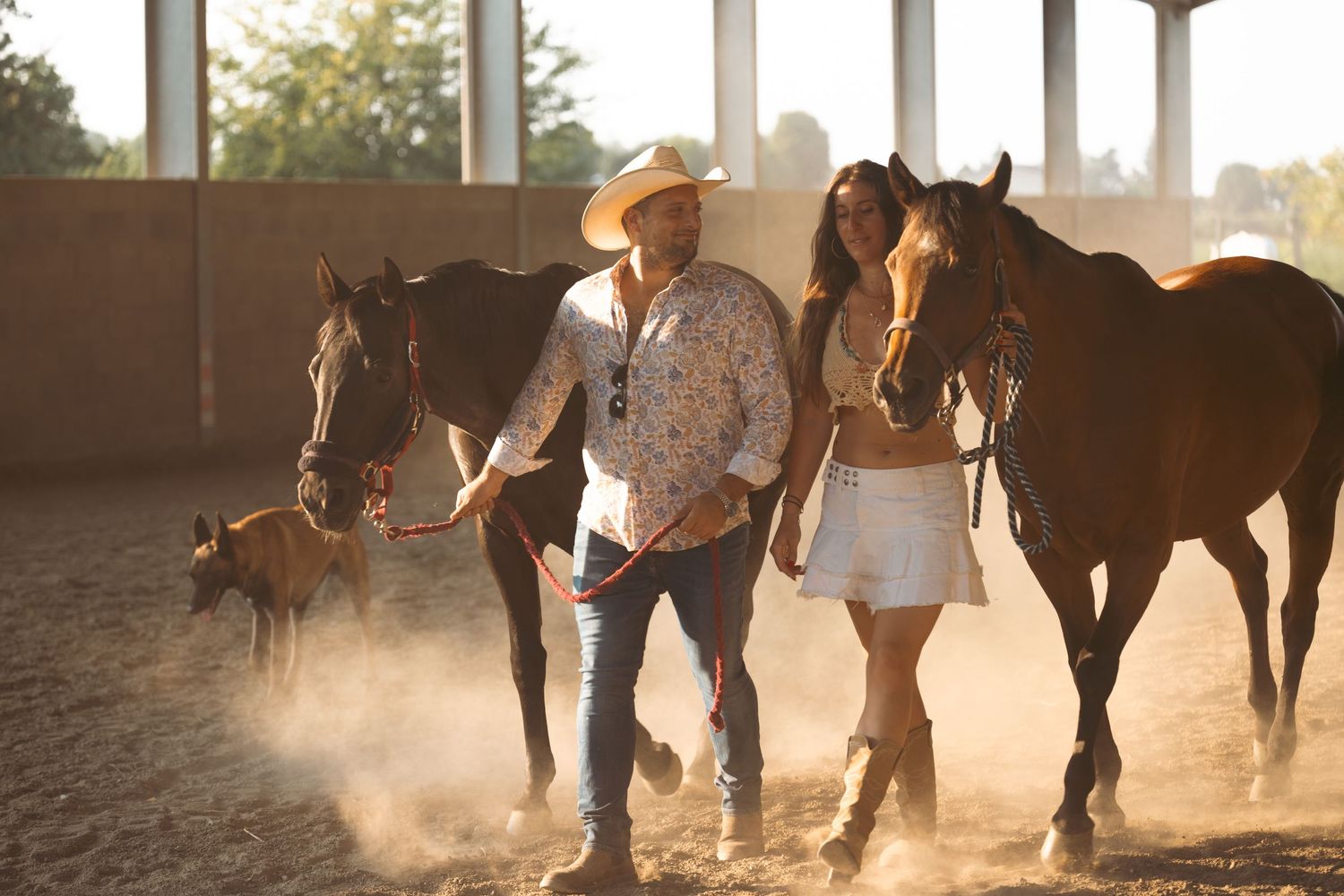 Horses and handlers creating dust clouds in a golden-lit arena at dusk.