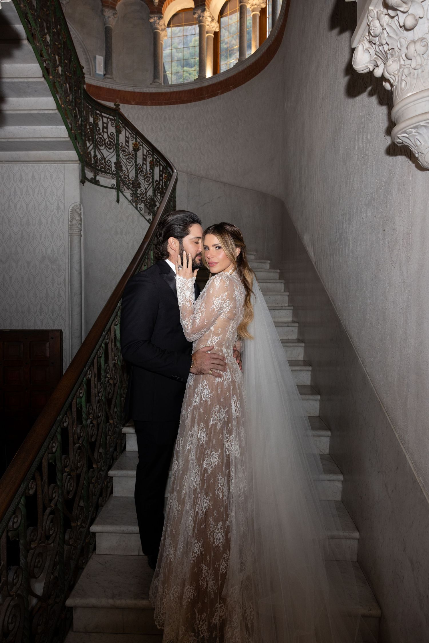 A romantic wedding couple embraces on an elegant staircase with ornate iron railings and classic architectural details.