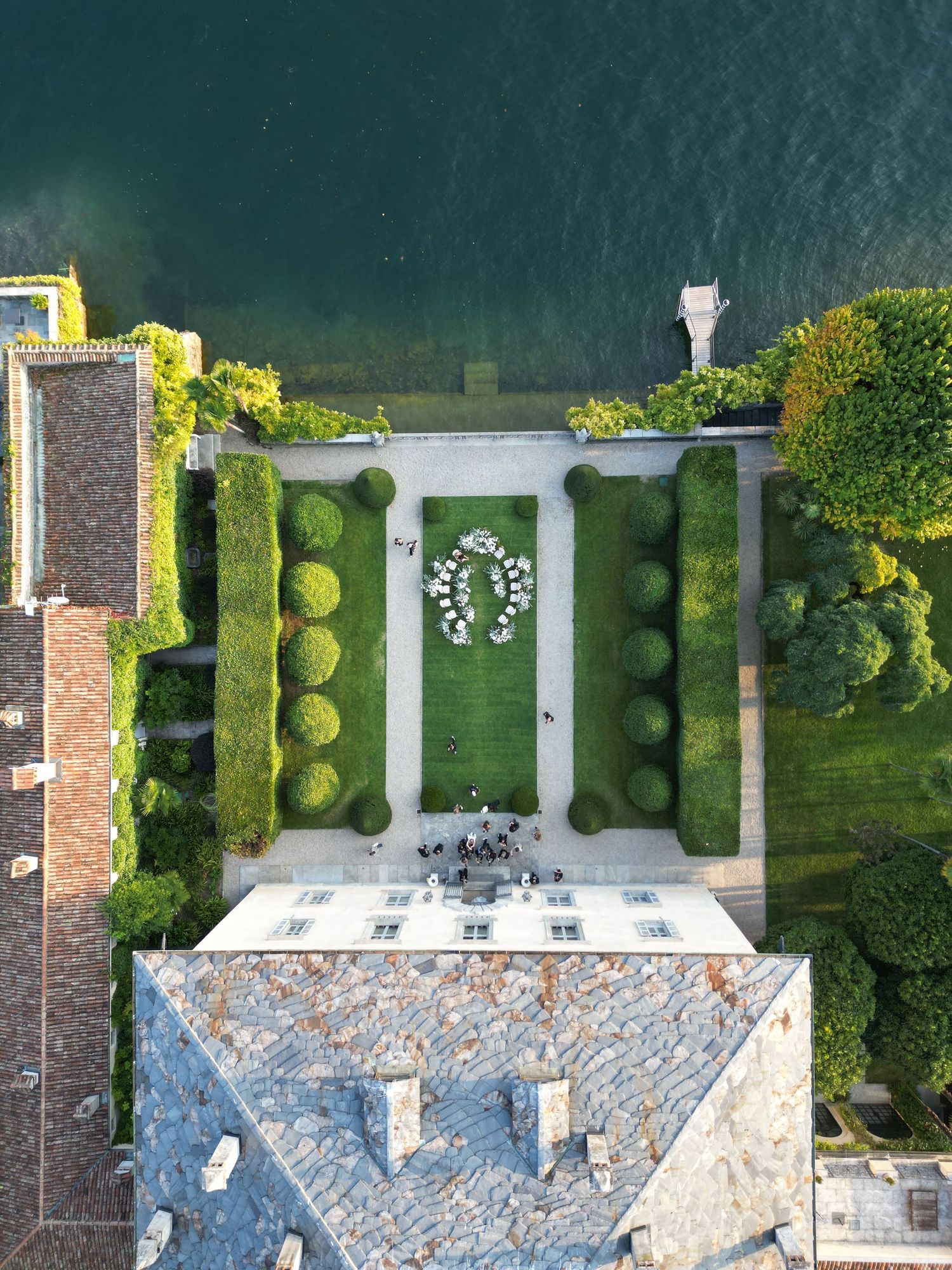 Aerial view of a formal garden with symmetrical hedges and a white floral arch arrangement seen from above a stone building.