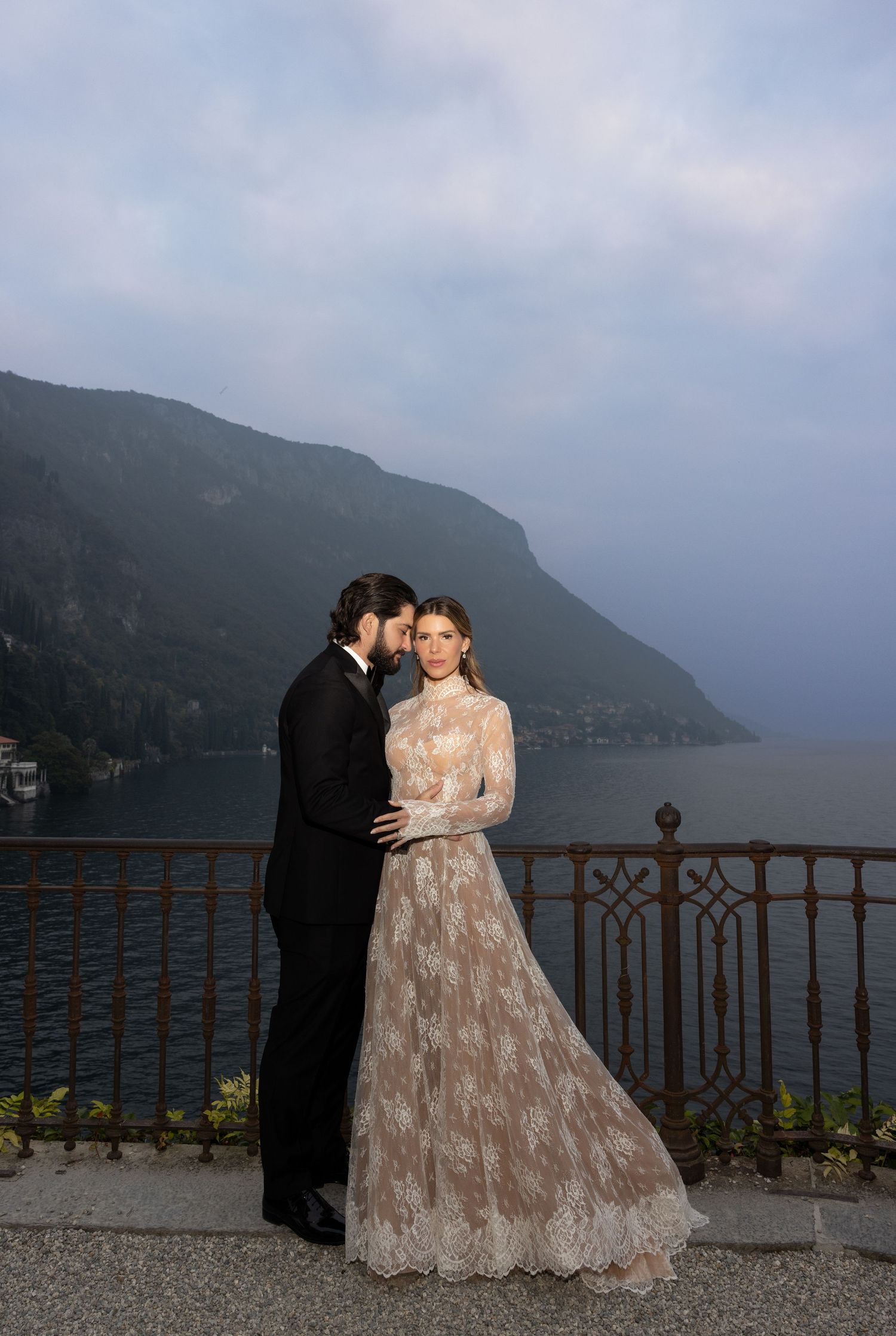 A couple in formal attire poses against a scenic mountain lake backdrop on a misty day.