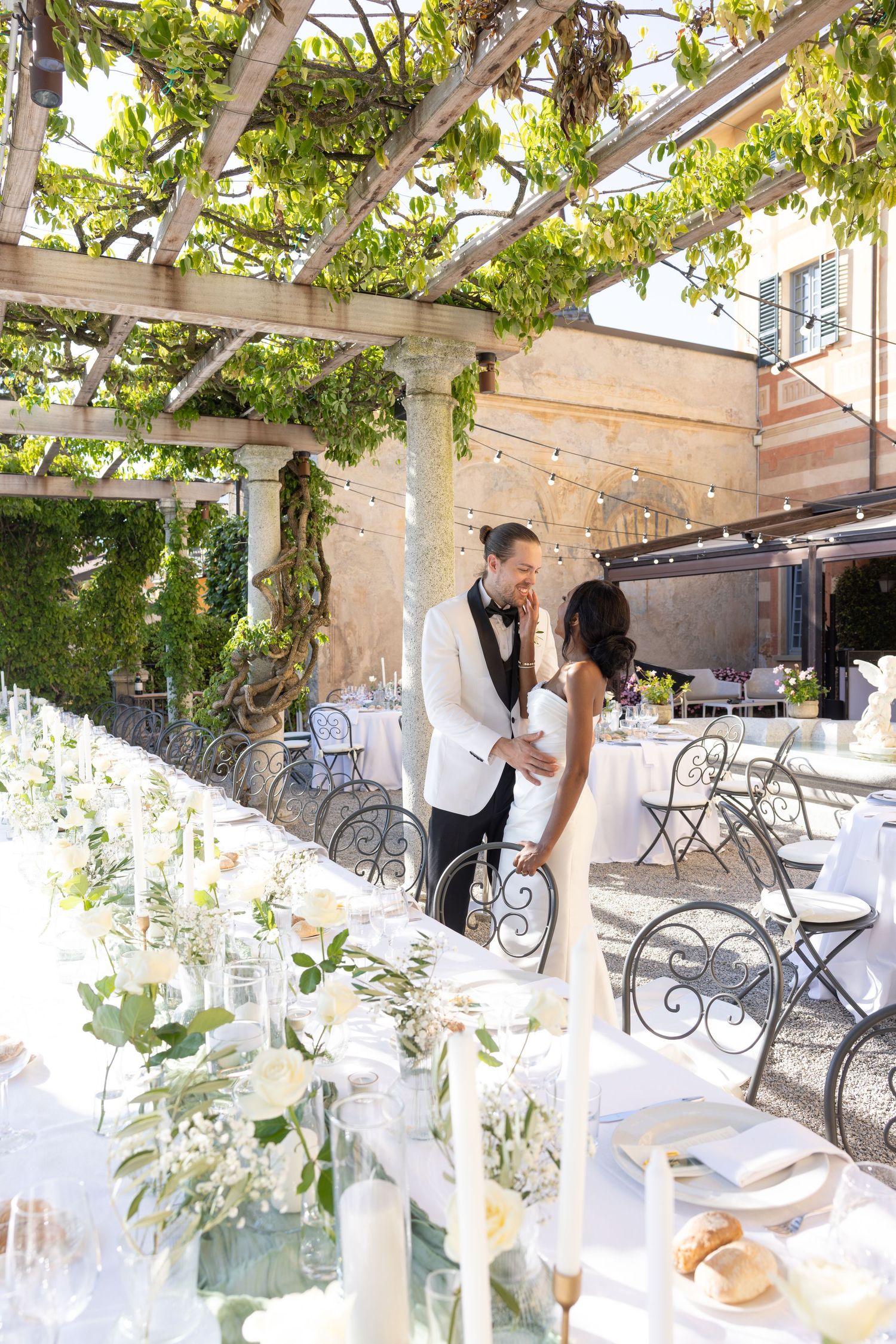 Couple sharing an intimate moment under a vine-covered pergola with elegant white decor and table settings.