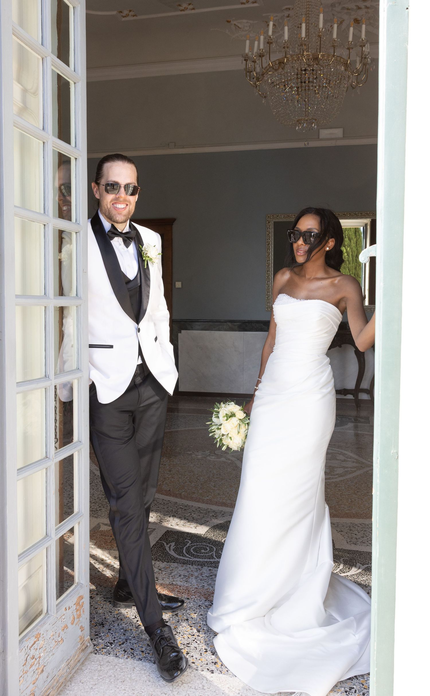 A bride in a white gown and groom in formal attire pose playfully in doorways at an elegant venue with chandeliers.