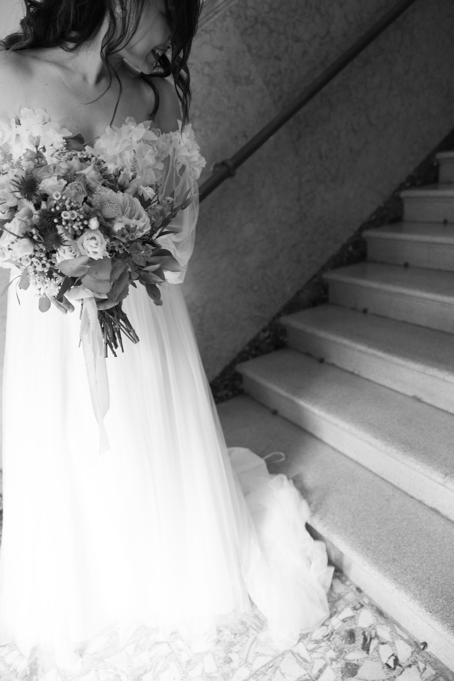 Black and white photo of bride holding elegant floral bouquet on staircase.