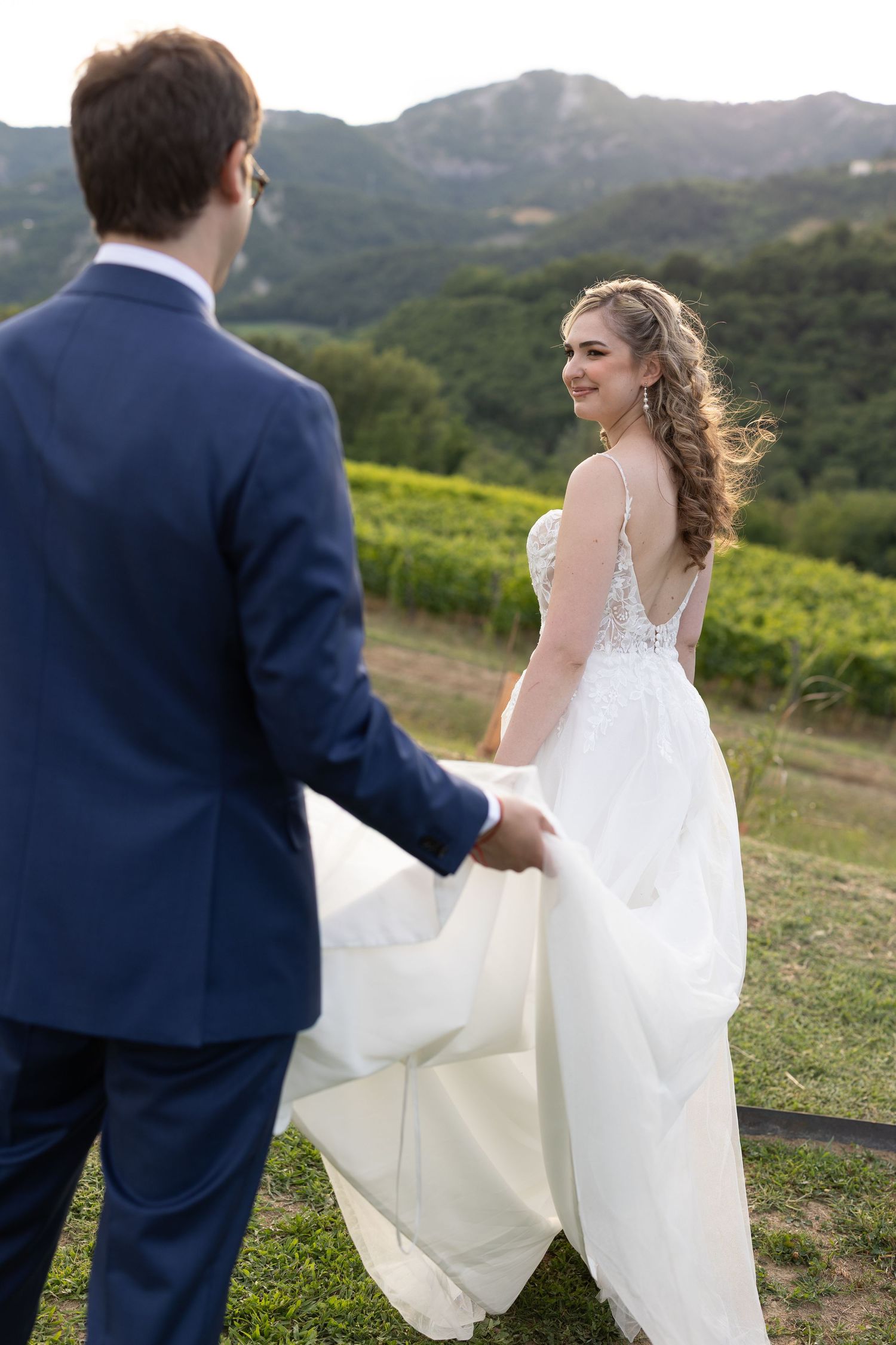 Wedding couple walking through scenic vineyard with mountains in background.