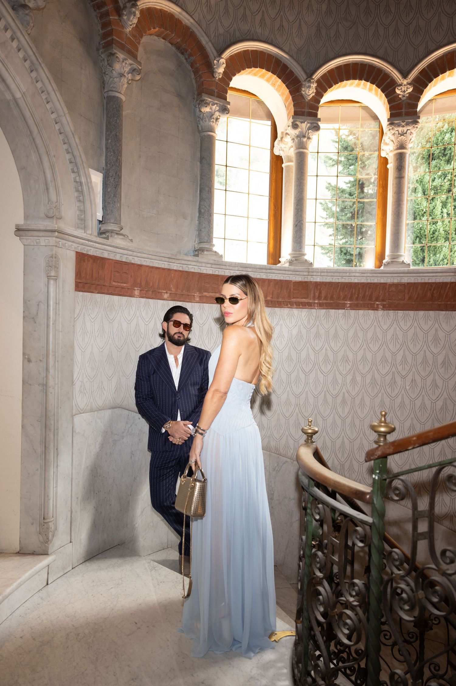 Romantic couple poses on elegant stone staircase with arched windows and ornate architectural details in castle interior.