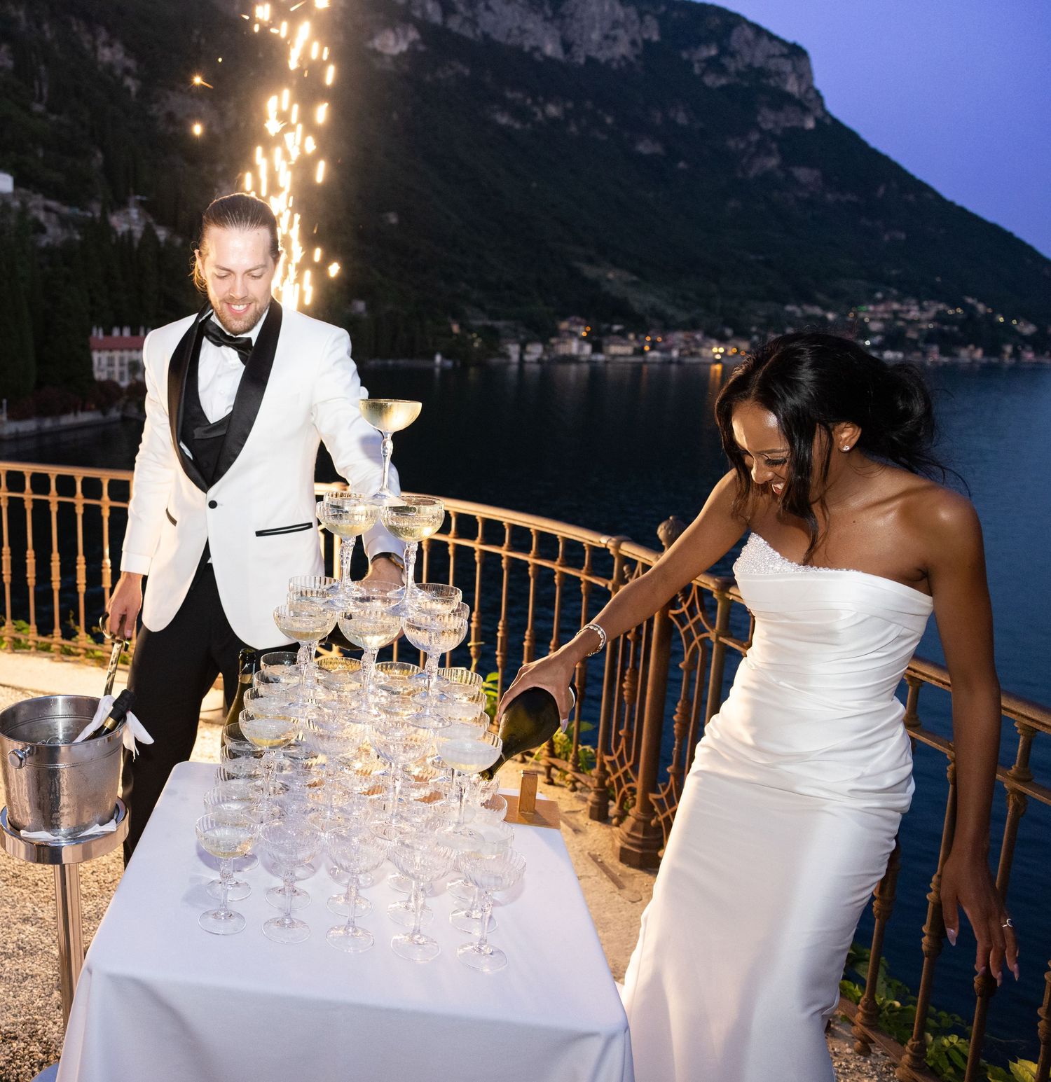Champagne tower being filled at an elegant outdoor wedding reception with coastal mountains in the background at dusk.
