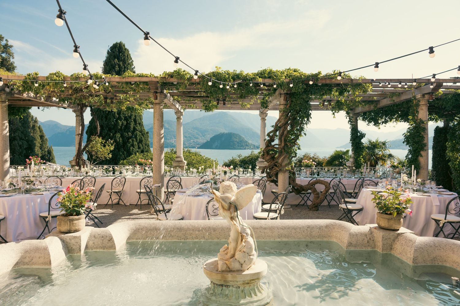 String lights illuminate an elegant outdoor wedding reception setup overlooking a scenic coastal view at sunset.