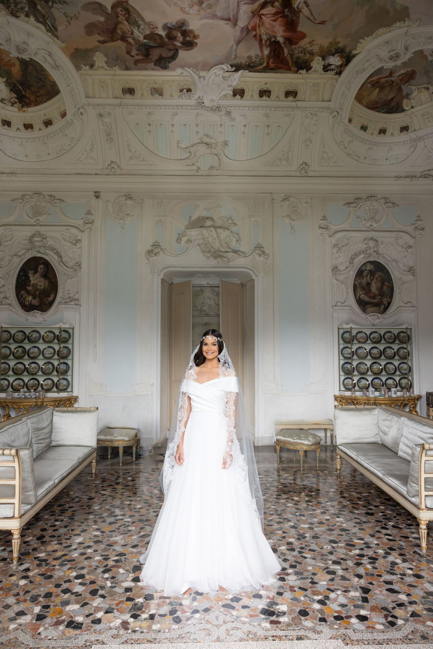A bride in a white wedding dress poses in an ornate room with decorative ceiling frescoes and patterned marble floors.