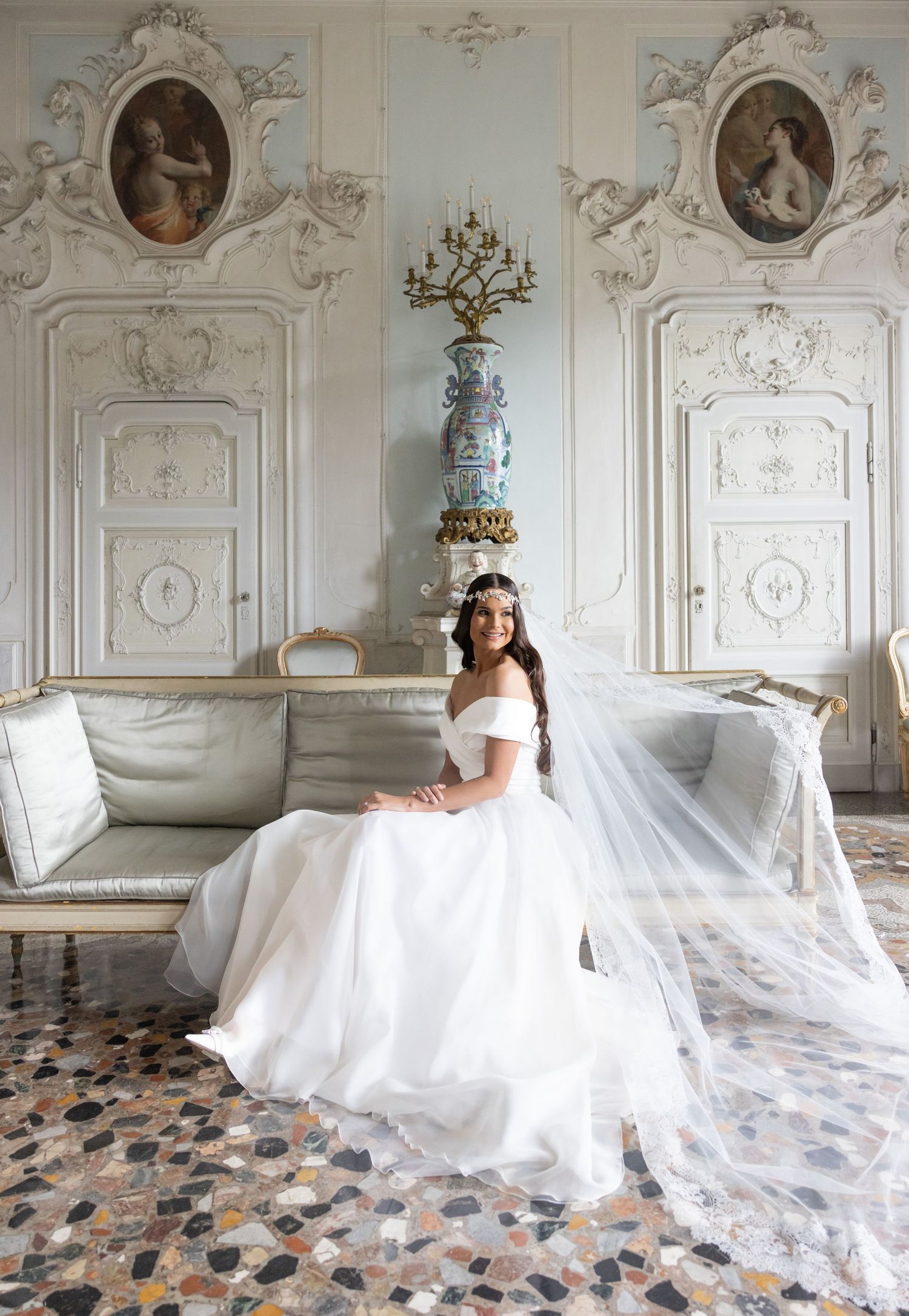 A bride in a flowing white gown poses on an antique sofa in an ornate room with painted wall panels.