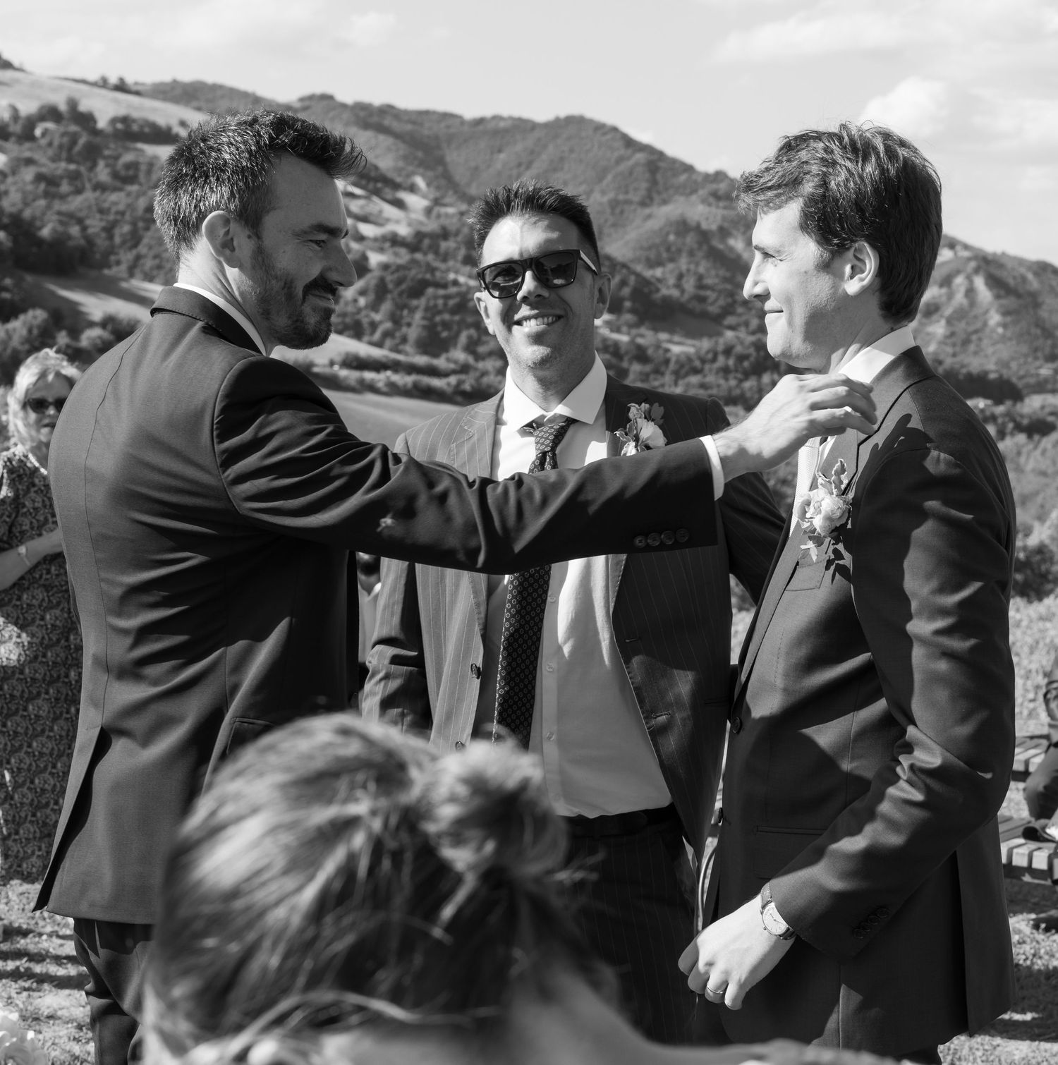 Three people in suits share a friendly moment at an outdoor mountain wedding.