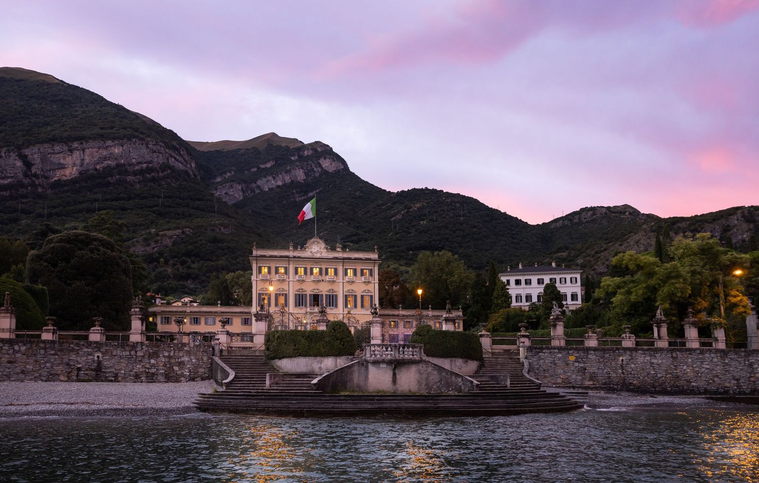 Lakeside villas and buildings at dusk with purple sky and mountain backdrop.