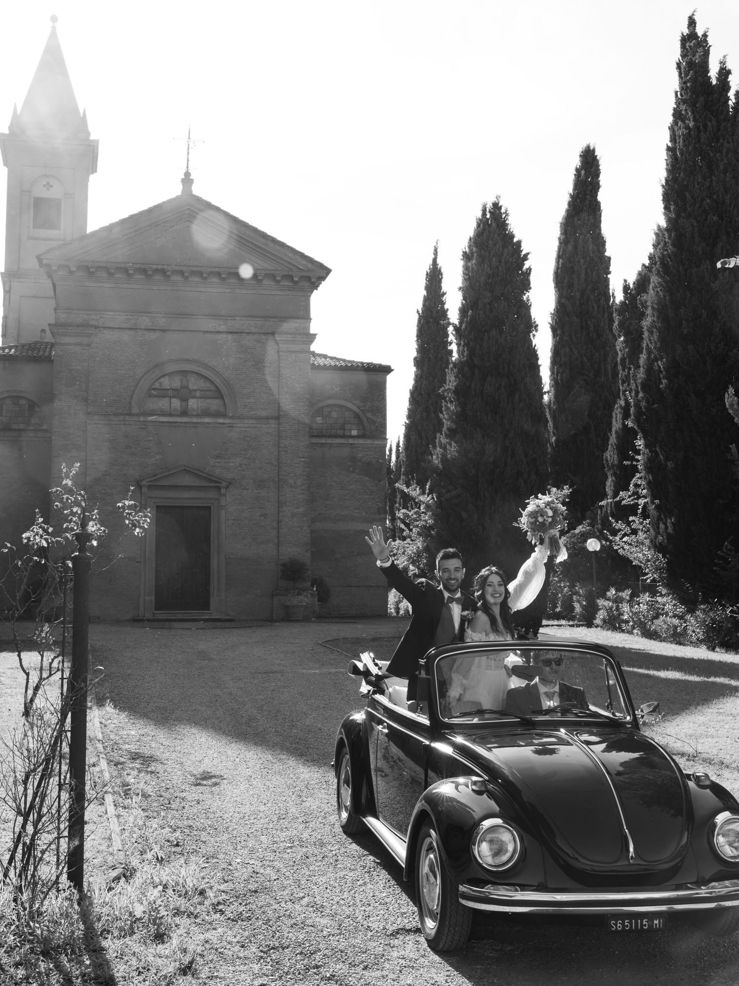 Vintage Volkswagen Beetle parked outside a historic church with cypress trees.