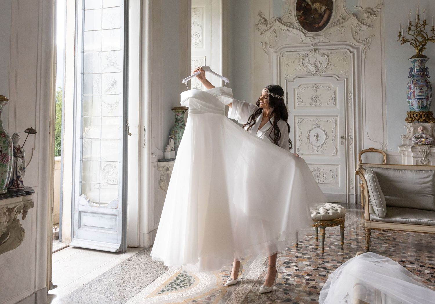 Elegant white wedding dress displayed in ornate room with decorative ceiling and large windows.