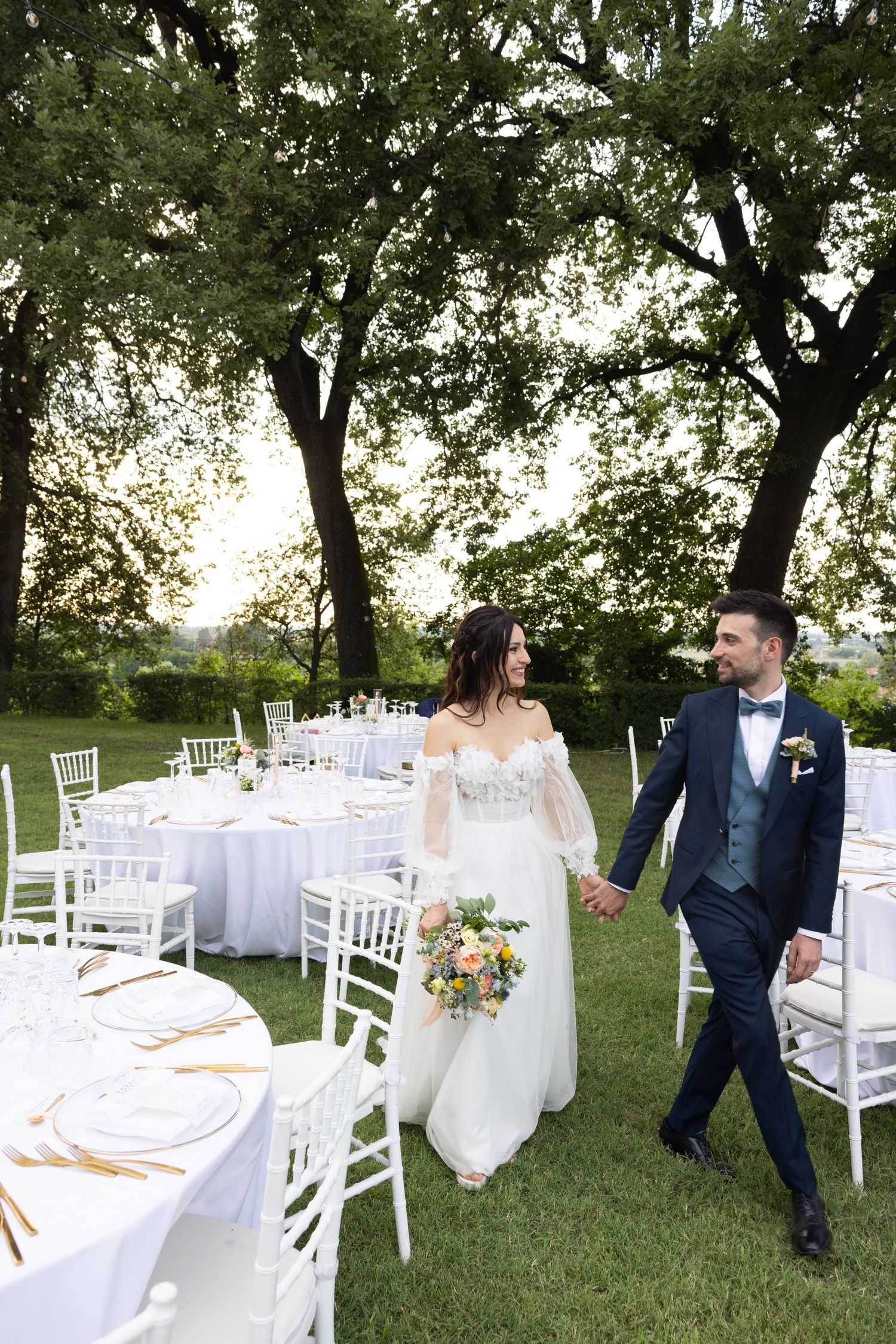 Outdoor wedding reception setup with white chairs and tables under tall trees.