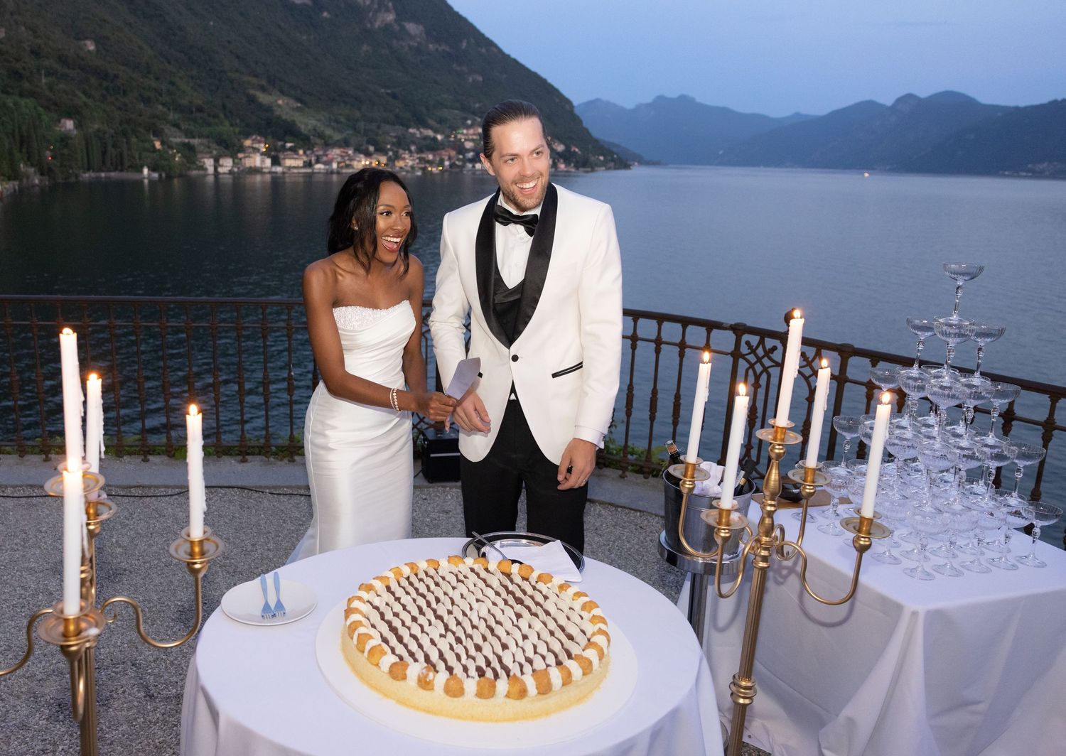 Wedding reception tables set with elegant white linens and candelabras along a scenic lakeside terrace at sunset.