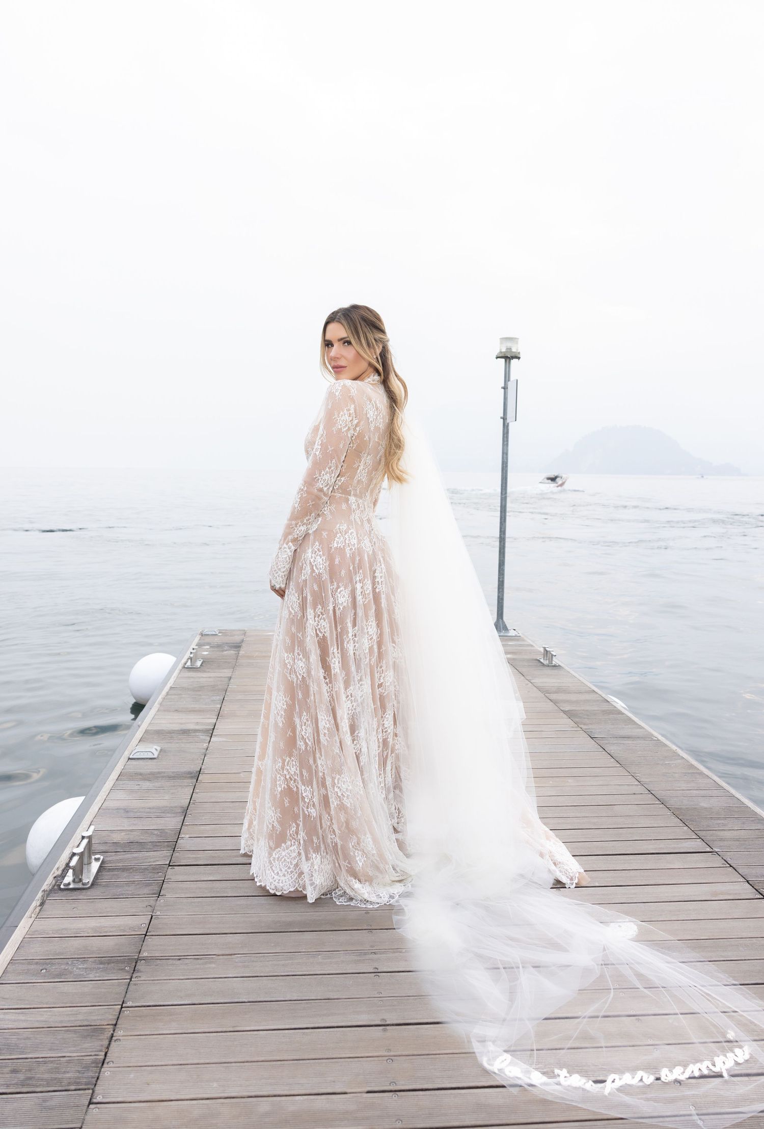 A bride in a flowing lace gown and long veil walks along a wooden pier by the ocean on her wedding day.