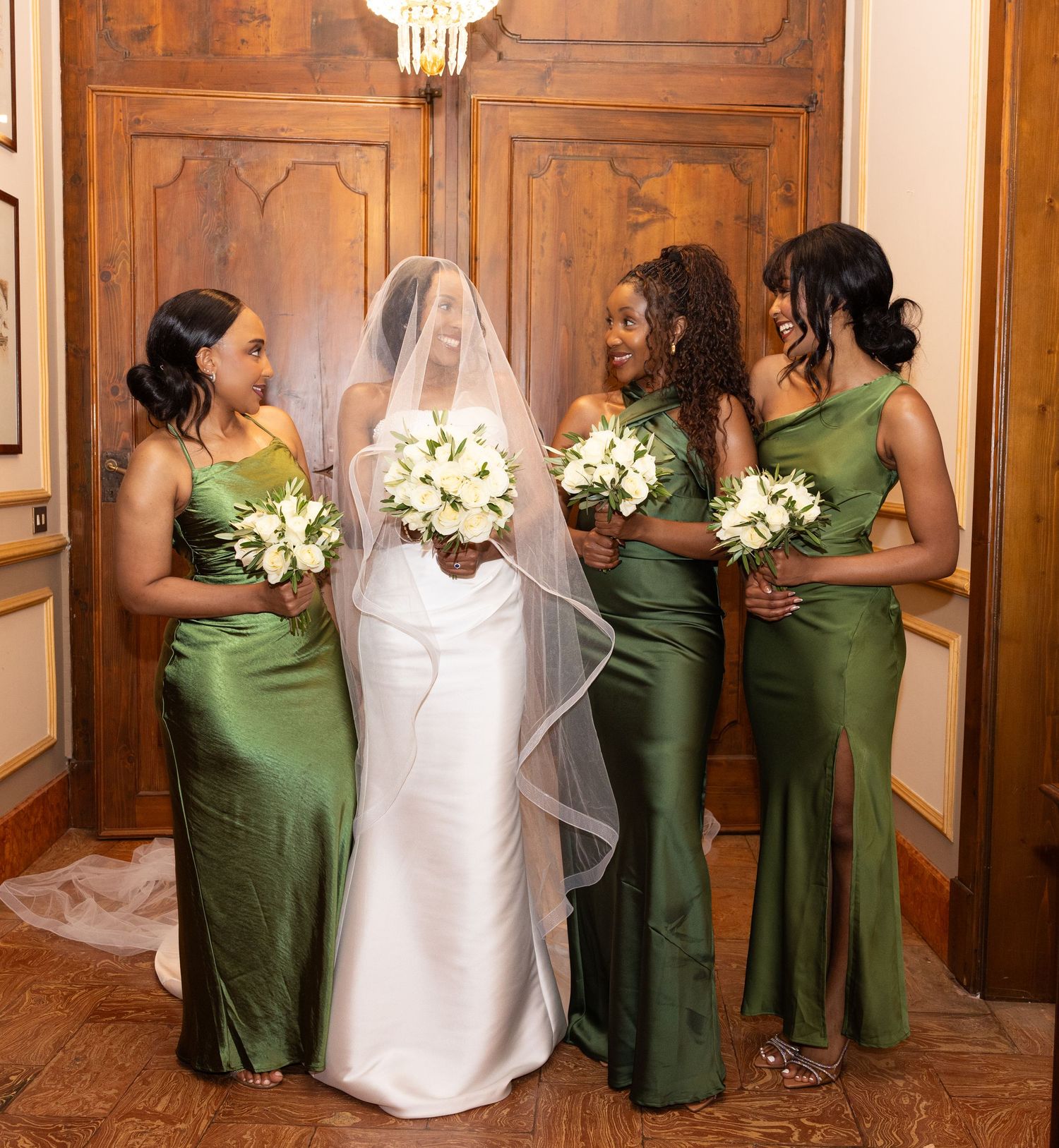 Bridesmaids in elegant green dresses pose with bride in white gown holding bouquets in ornate wooden hallway.