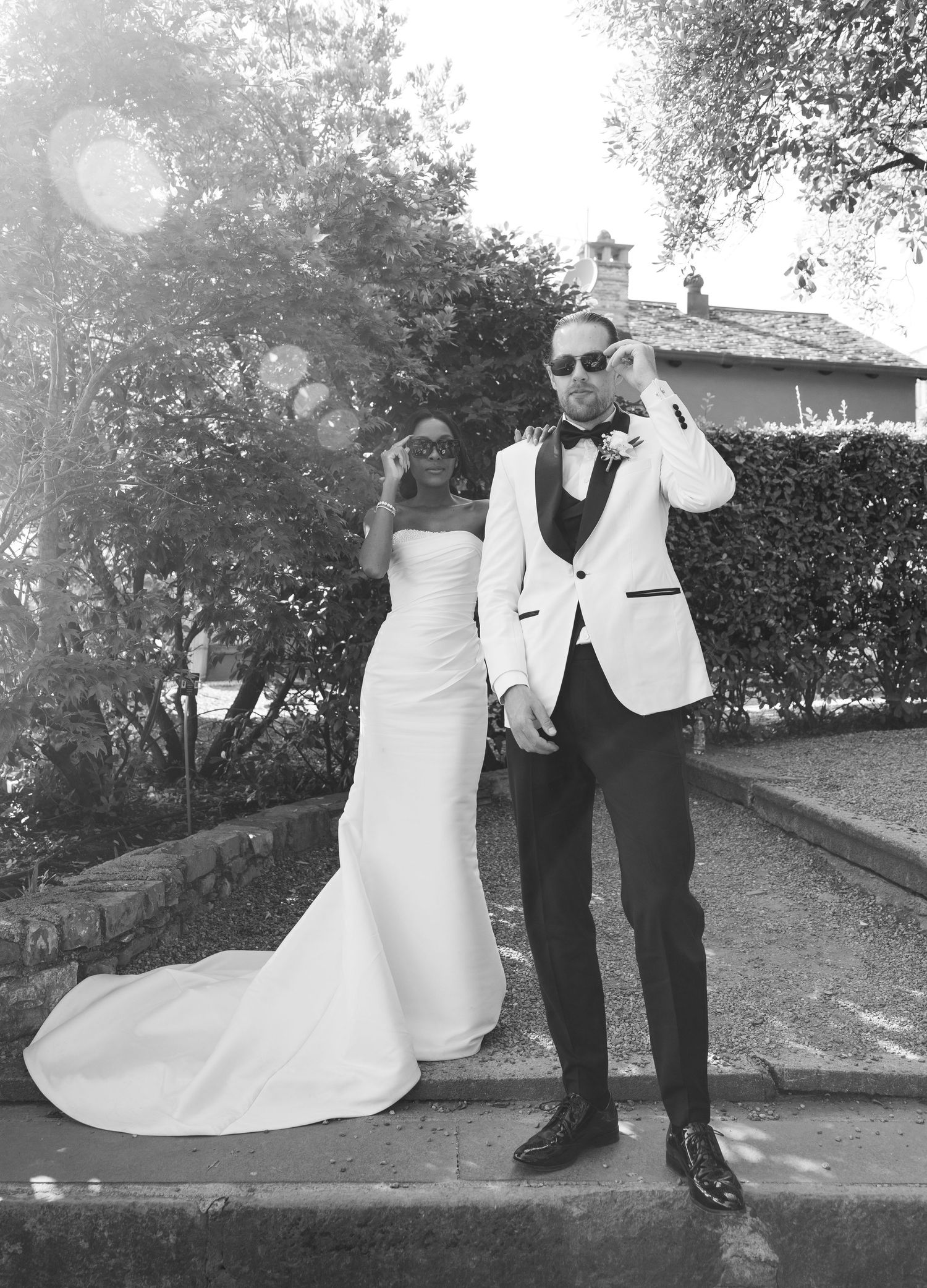 A couple poses together outdoors in formal wedding attire, with the bride in white and groom in a light-colored jacket.