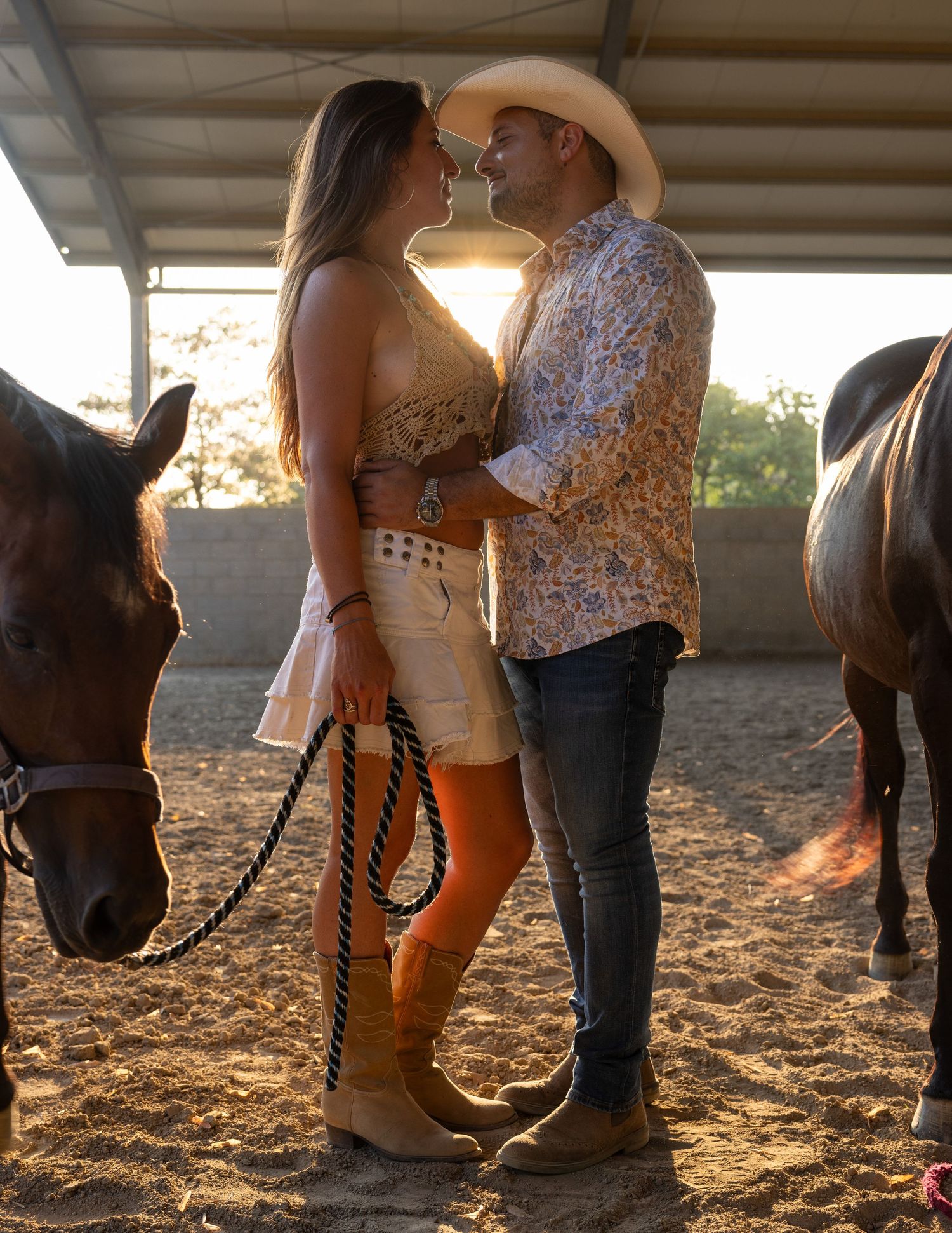 People in western wear interacting with horses in a covered arena at sunset.