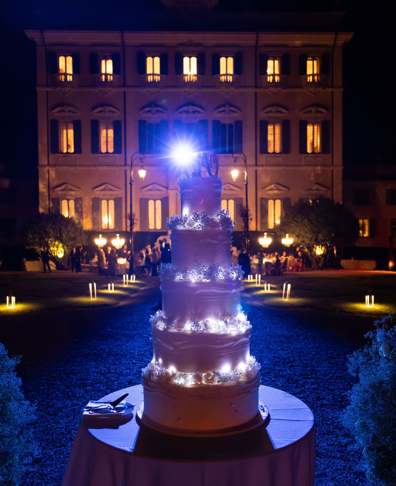 Elegant wedding cake illuminated at night with a grand mansion venue in the background.