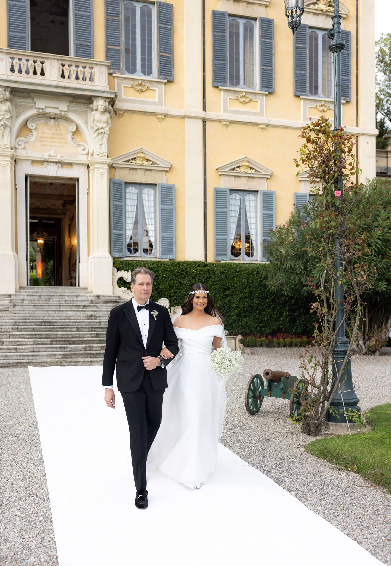 Wedding couple walking along a grand yellow Italian villa with stone steps and blue shutters.