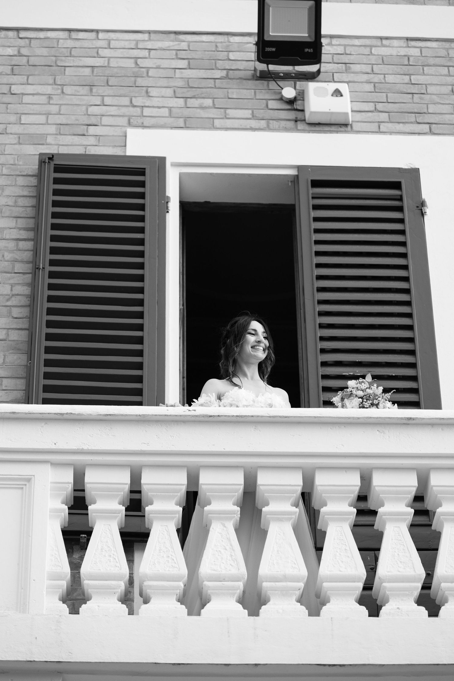 A black and white photo showing a balcony with white railings and shuttered windows on a brick building.