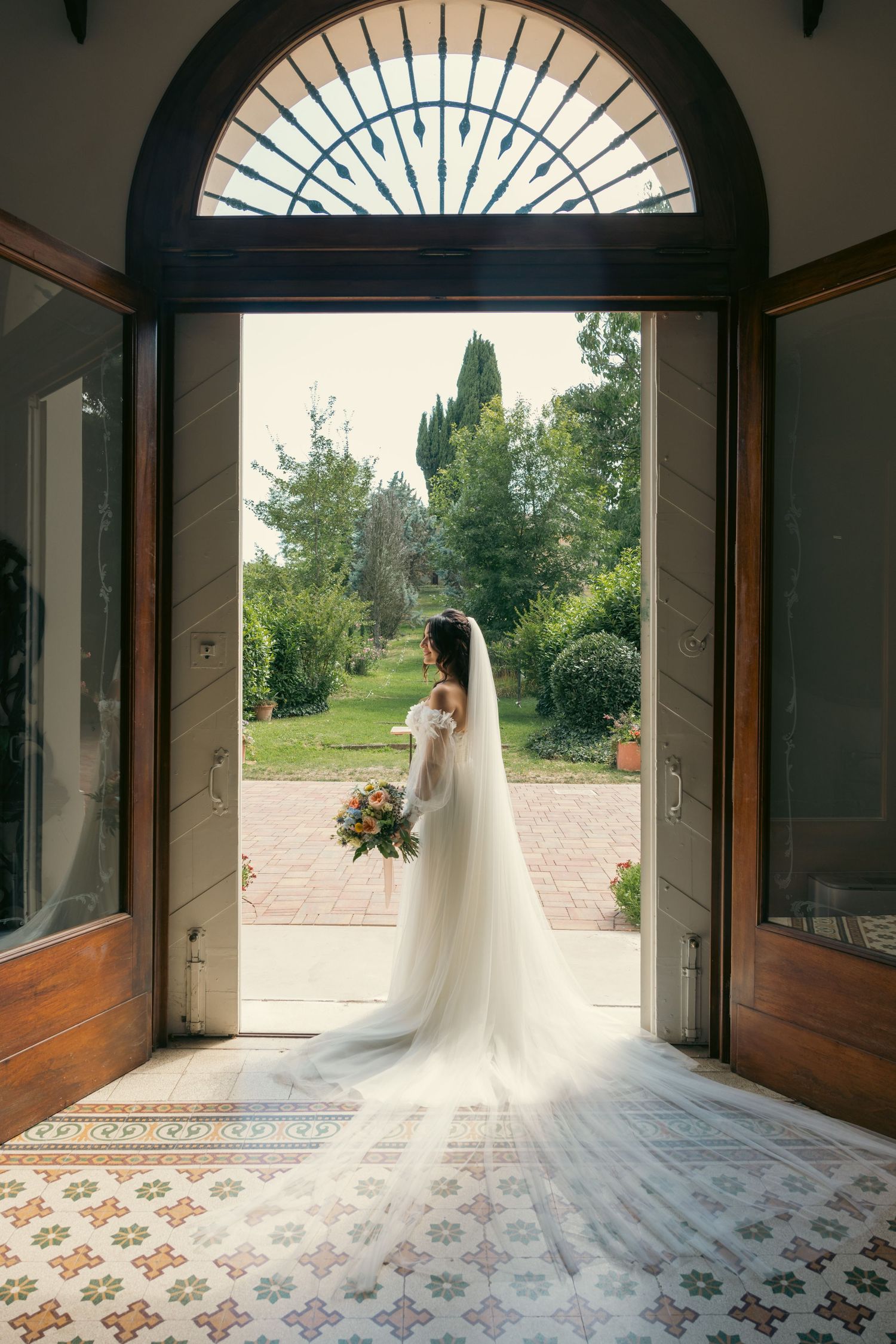 Sunlight streams through an arched doorway, illuminating a bridal silhouette on patterned floor tiles.