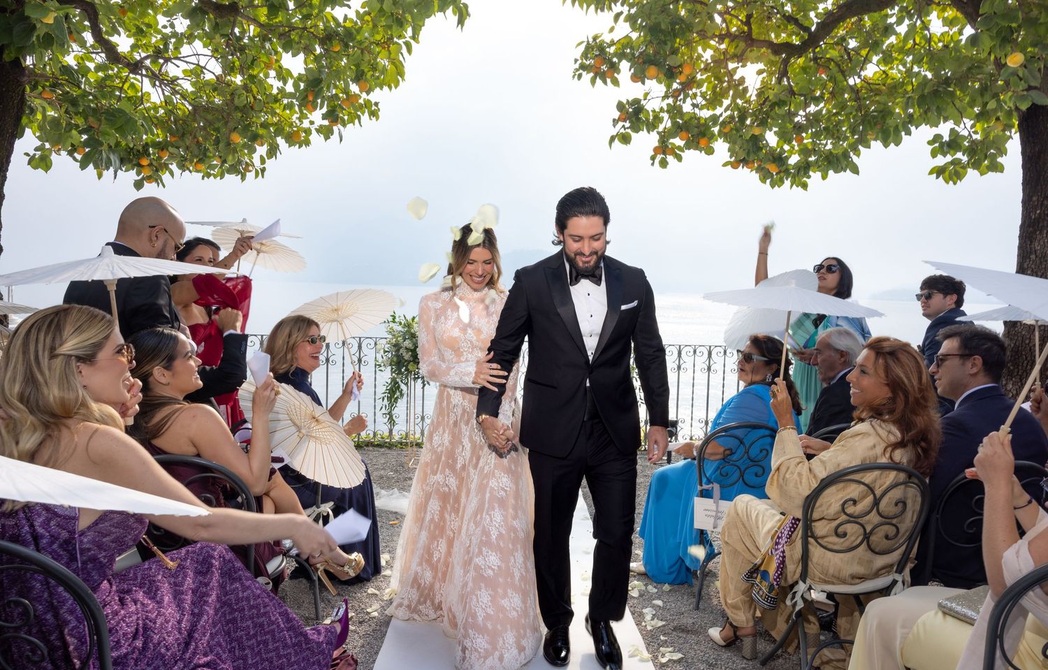 Wedding celebration captured outdoors with guests gathering under green trees.
