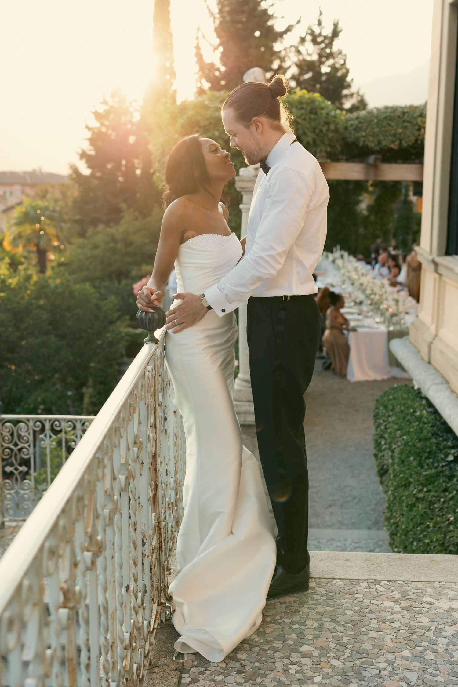 Romantic wedding couple embraces on a stone balcony at sunset with garden views in the background.