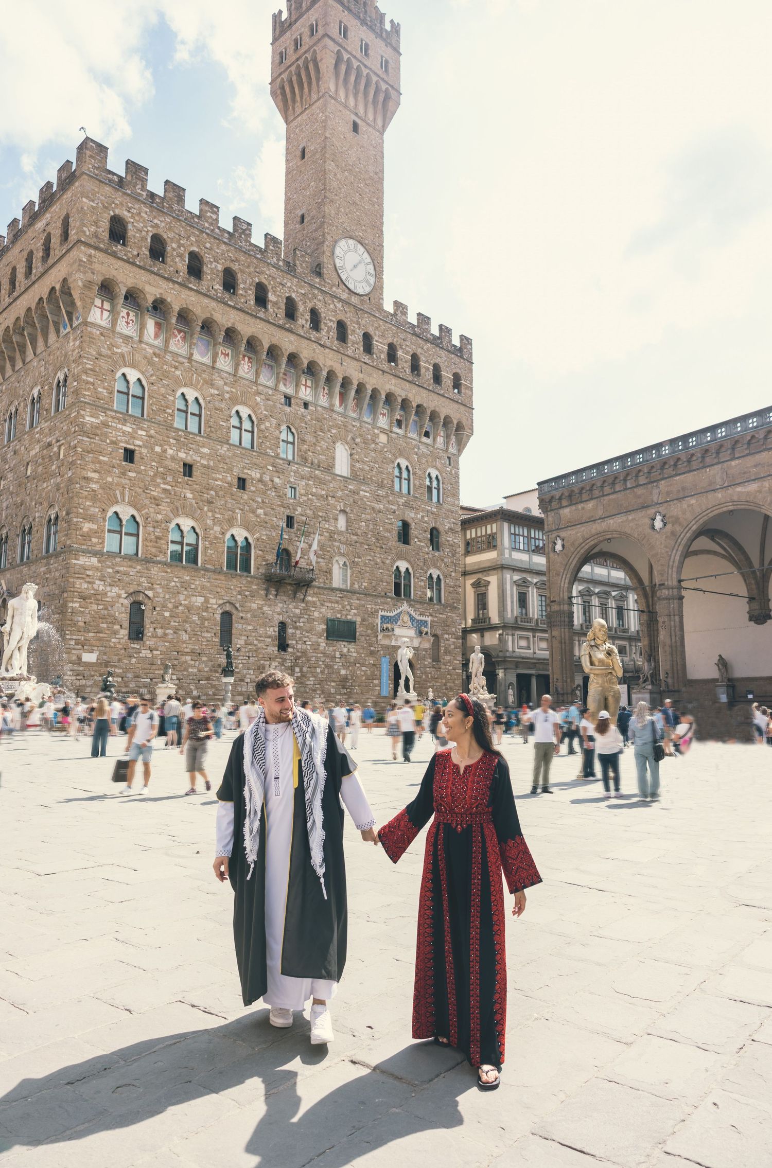 Tourists in traditional dress stroll through Piazza della Signoria in Florence with Palazzo Vecchio tower in background.