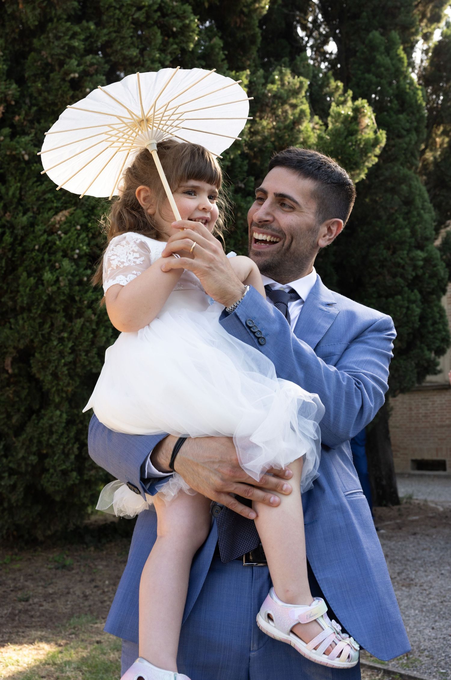 Joyful moment at a wedding with a white parasol and blue suit against a cypress tree backdrop.