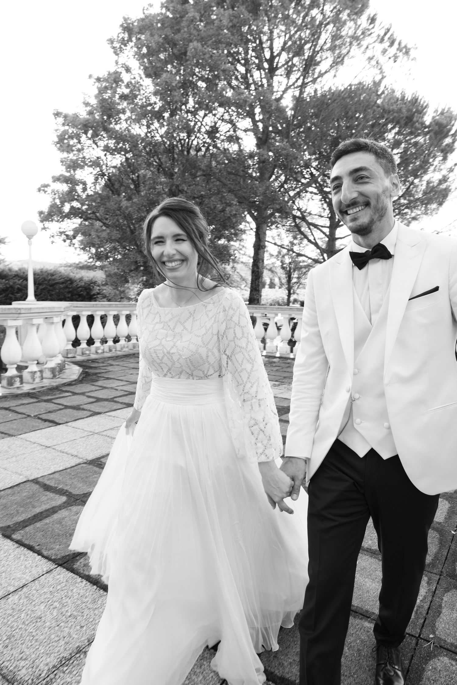Black and white photo of happy newlyweds walking hand in hand on stone terrace.