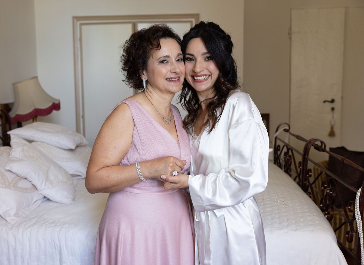 Bridesmaids in pink and white robes share joyful moments on a bed during wedding preparations.
