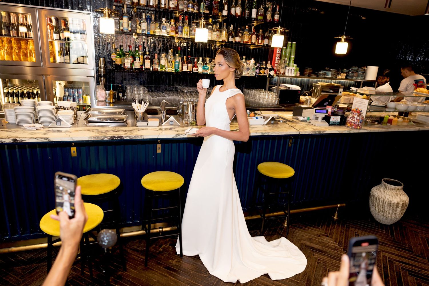Elegant bar setting with backlit liquor shelves and yellow barstools displaying a white wedding dress.