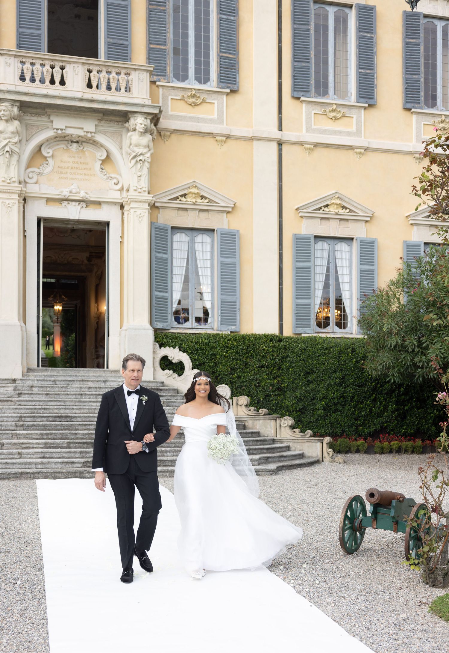 A wedding couple walks along a grand Italian villa's exterior with classic architecture and stone steps.