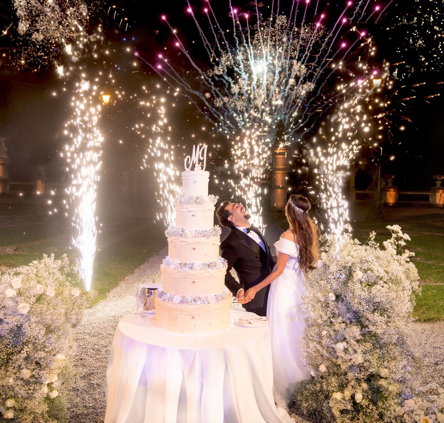 Wedding cake cutting moment captured with spectacular firework fountains in the background.