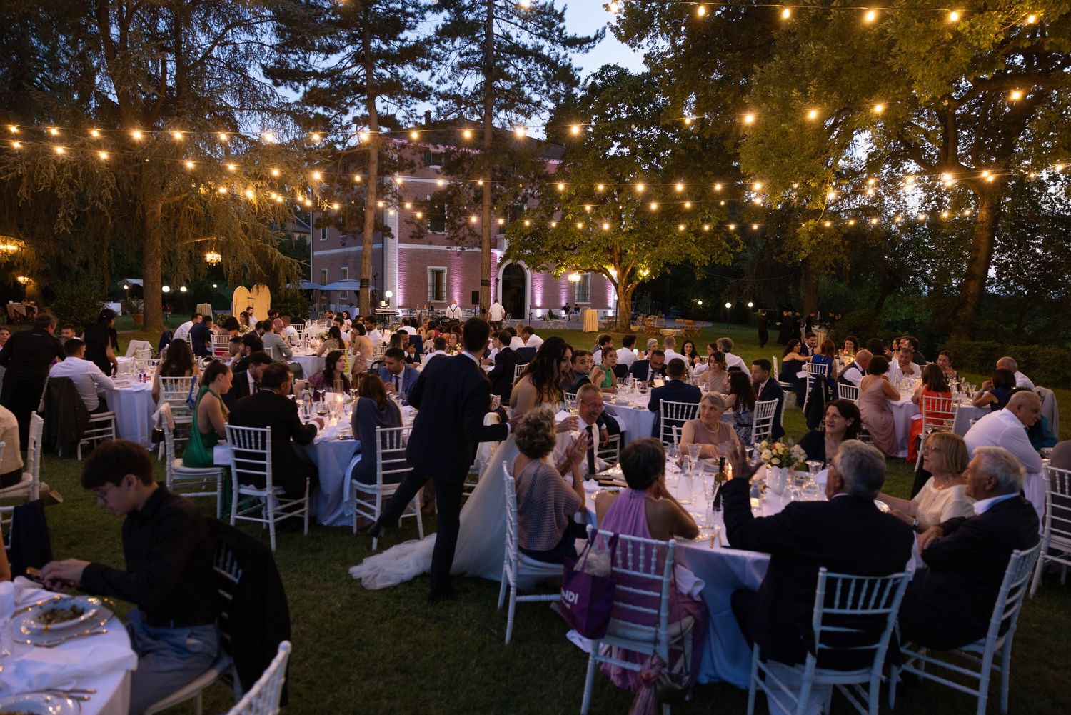 Outdoor evening wedding reception with string lights illuminating tables and guests.