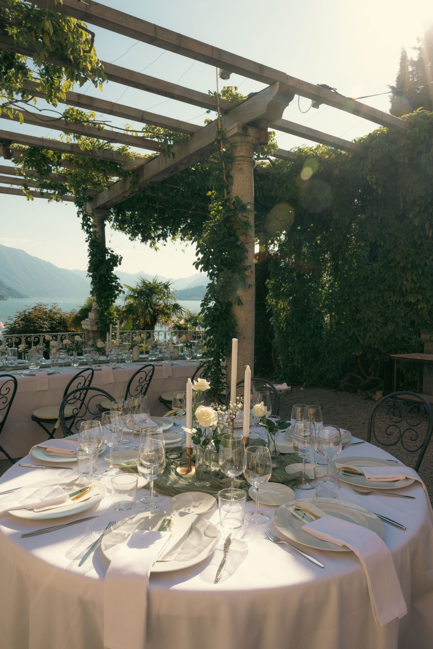 Elegant outdoor dining tables set with white linens and glassware under a vine-covered pergola overlooking a scenic coastline.