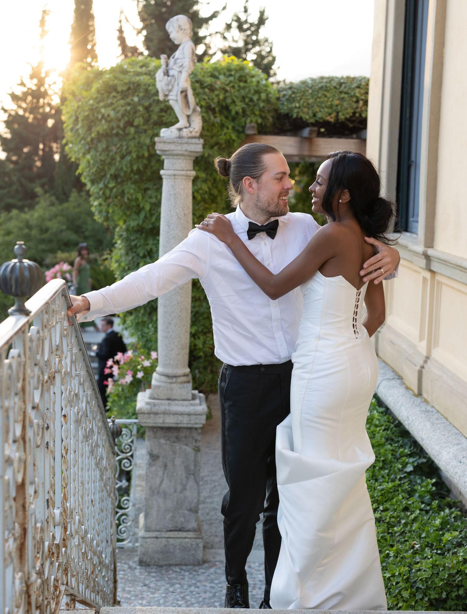 A romantic wedding moment captured at sunset on an elegant balcony with a classical stone statue in the background.