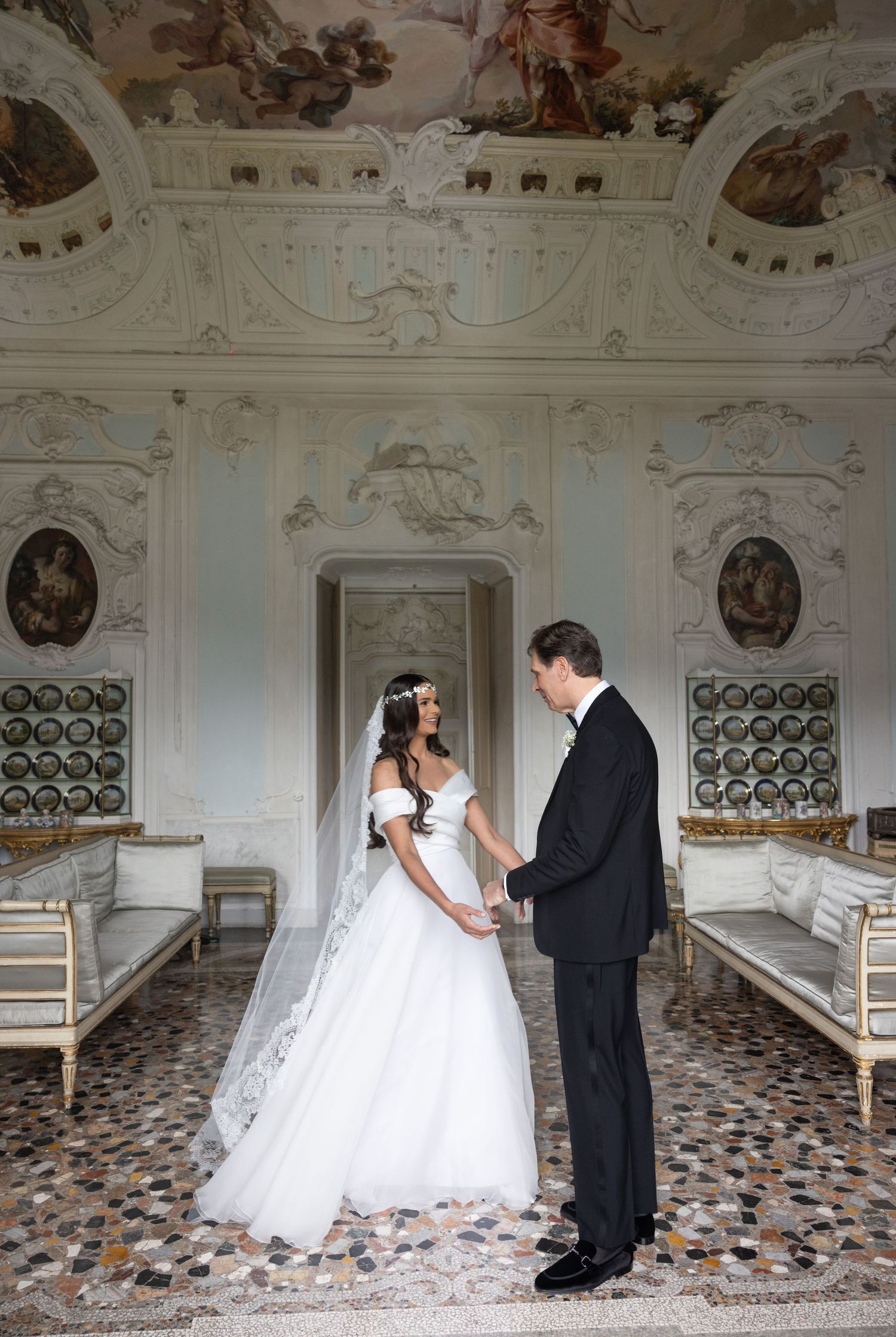 A bride and groom pose together in a historic venue with ornate white walls and painted ceilings.