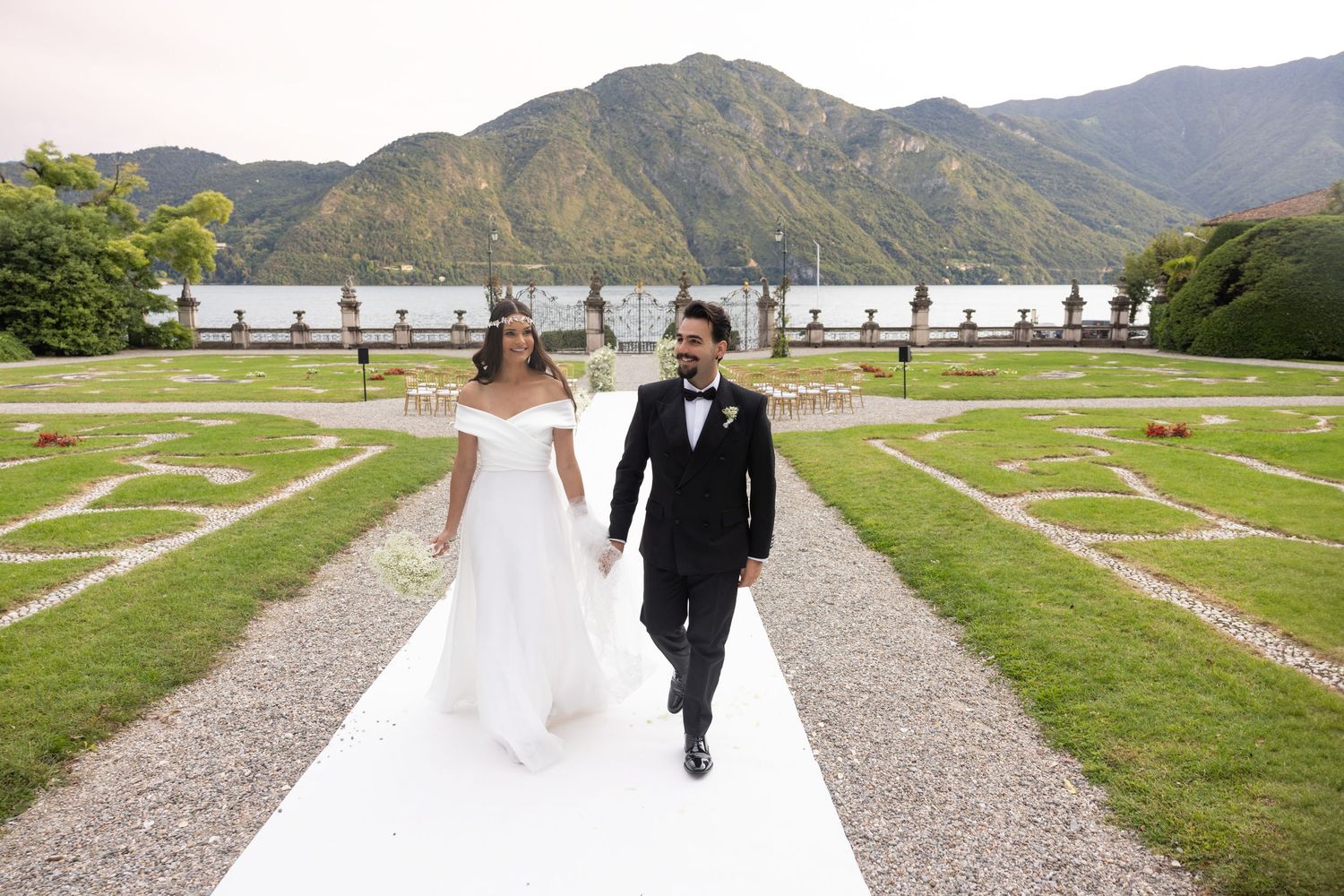 A couple strolls hand in hand on a gravel path with mountains and lake in the background.