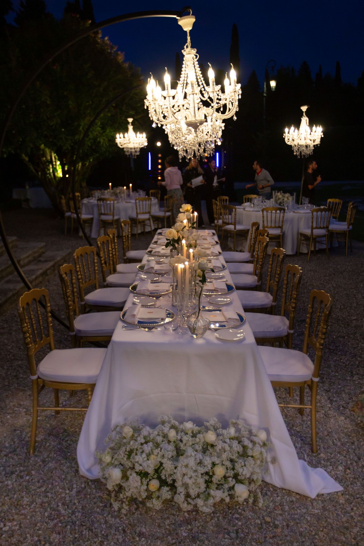 Elegant outdoor wedding reception tables with crystal chandeliers and white linens at night.