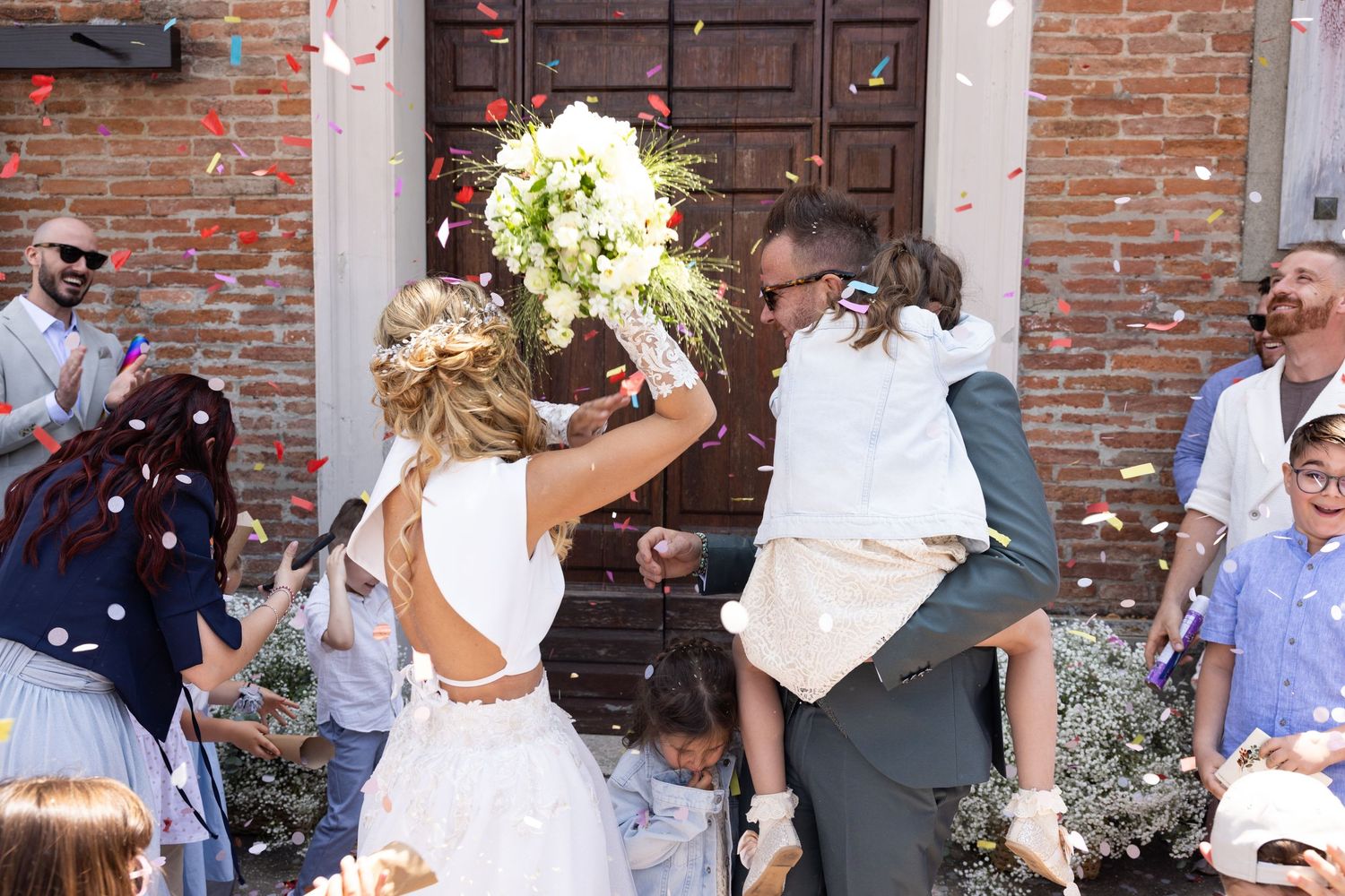 Wedding party exits through brick doorway as guests throw confetti in celebration.