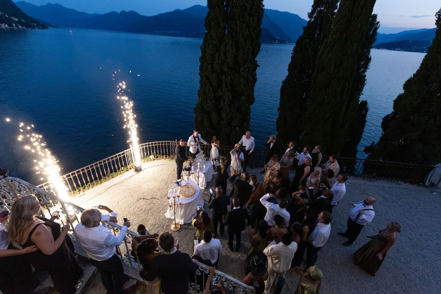 Wedding guests gather at dusk on a scenic lakeside terrace with cypress trees and mountain views in Italy.