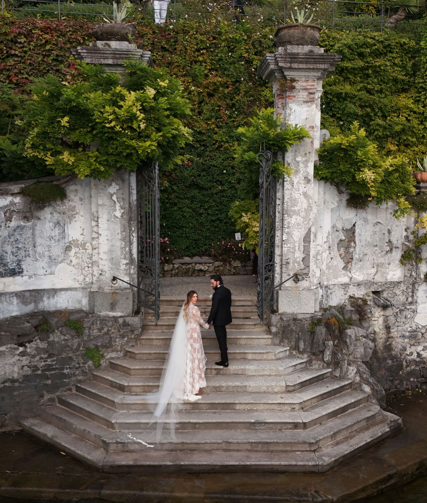 The Bride in a flowy dress poses gracefully on stone steps surrounded by ivy-covered walls in a garden setting.