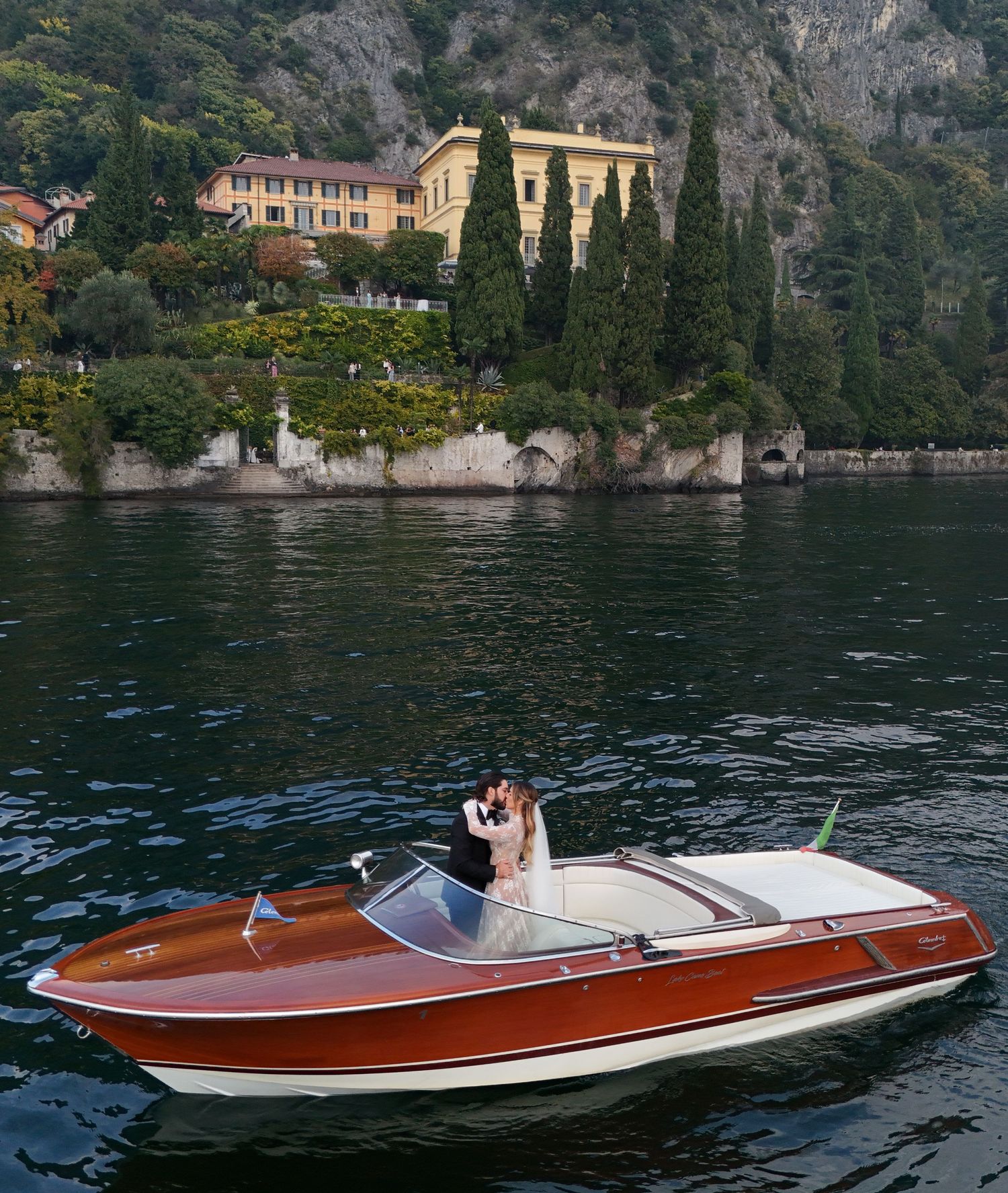 Vintage wooden boat cruising across Lake Como with mountains and Italian villas in the background.