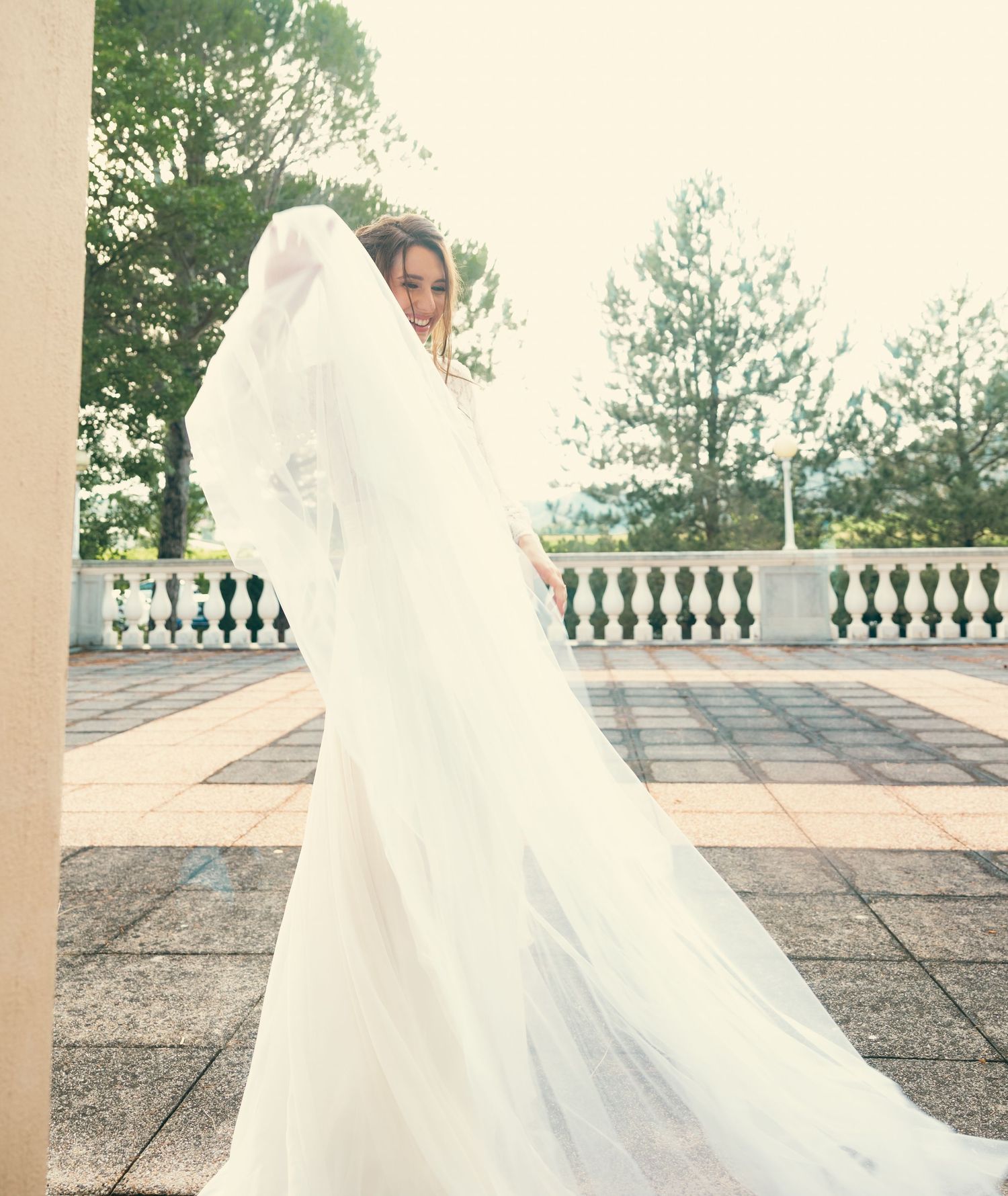 Bride in flowing white gown and veil poses on stone terrace with dramatic train.