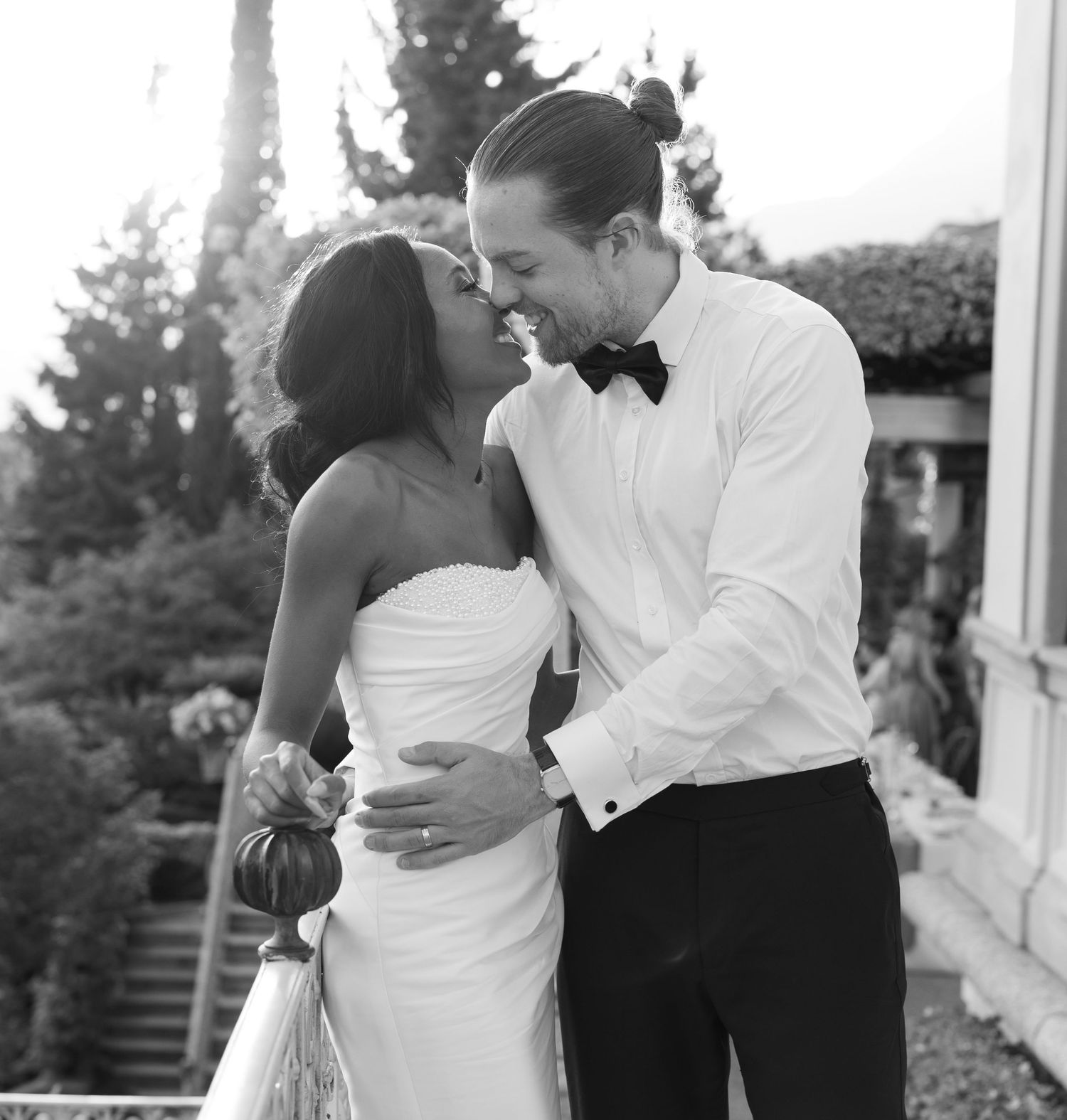 A romantic black and white wedding photo showing a couple embracing on a balcony with trees in the background.