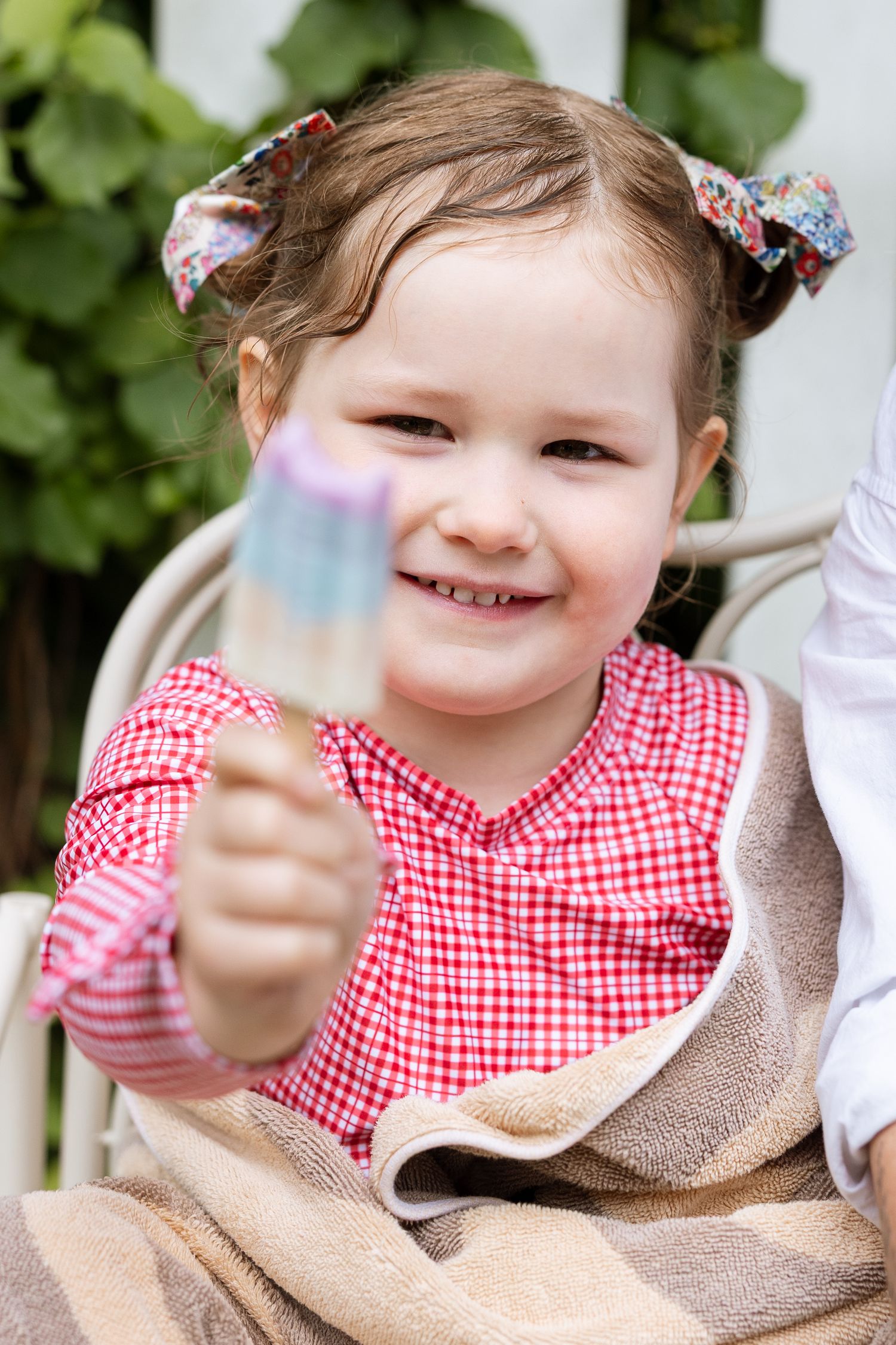 Child in red gingham dress wrapped in beige blanket smiles while holding iridescent object outdoors.